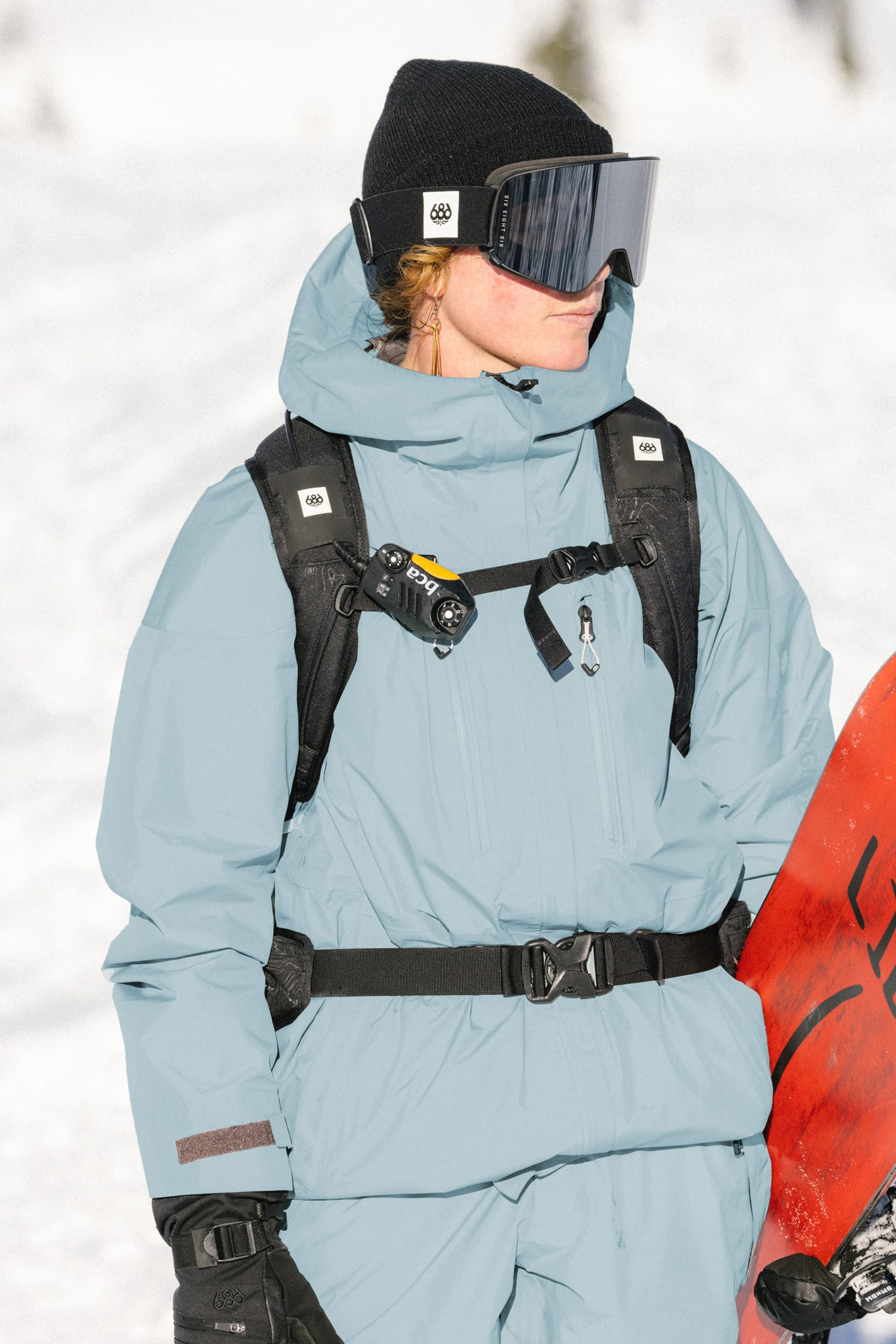 Wearing a 686 Women's GORE-TEX Skyline Thermagraph® Jacket in light blue, black gloves, a black beanie, ski goggles, and a black backpack with a camera on the strap, a person stands in the snow holding a red snowboard.