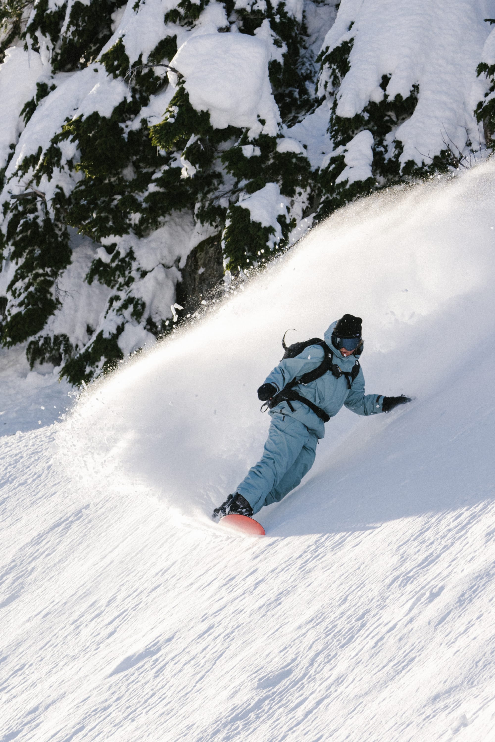A snowboarder wears the 686 Women's GORE-TEX Skyline Thermagraph® Jacket, gliding down a snowy slope and kicking up powder, with snow-covered evergreens in the background.