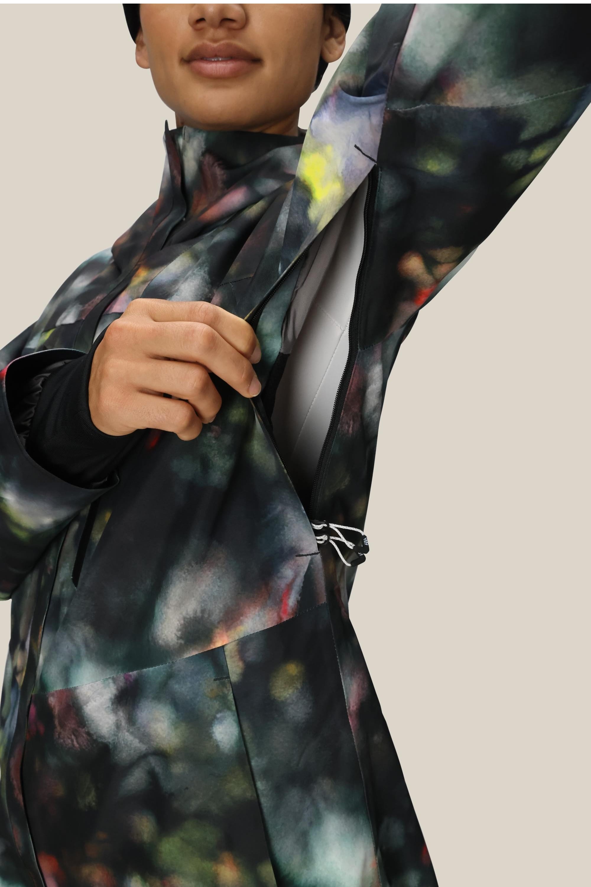 A woman unzips the side vent of a 686 Women's GORE-TEX Skyline Thermagraph® Jacket, revealing an inner layer. The image is cropped to show just her lower face and upper body against a plain background.