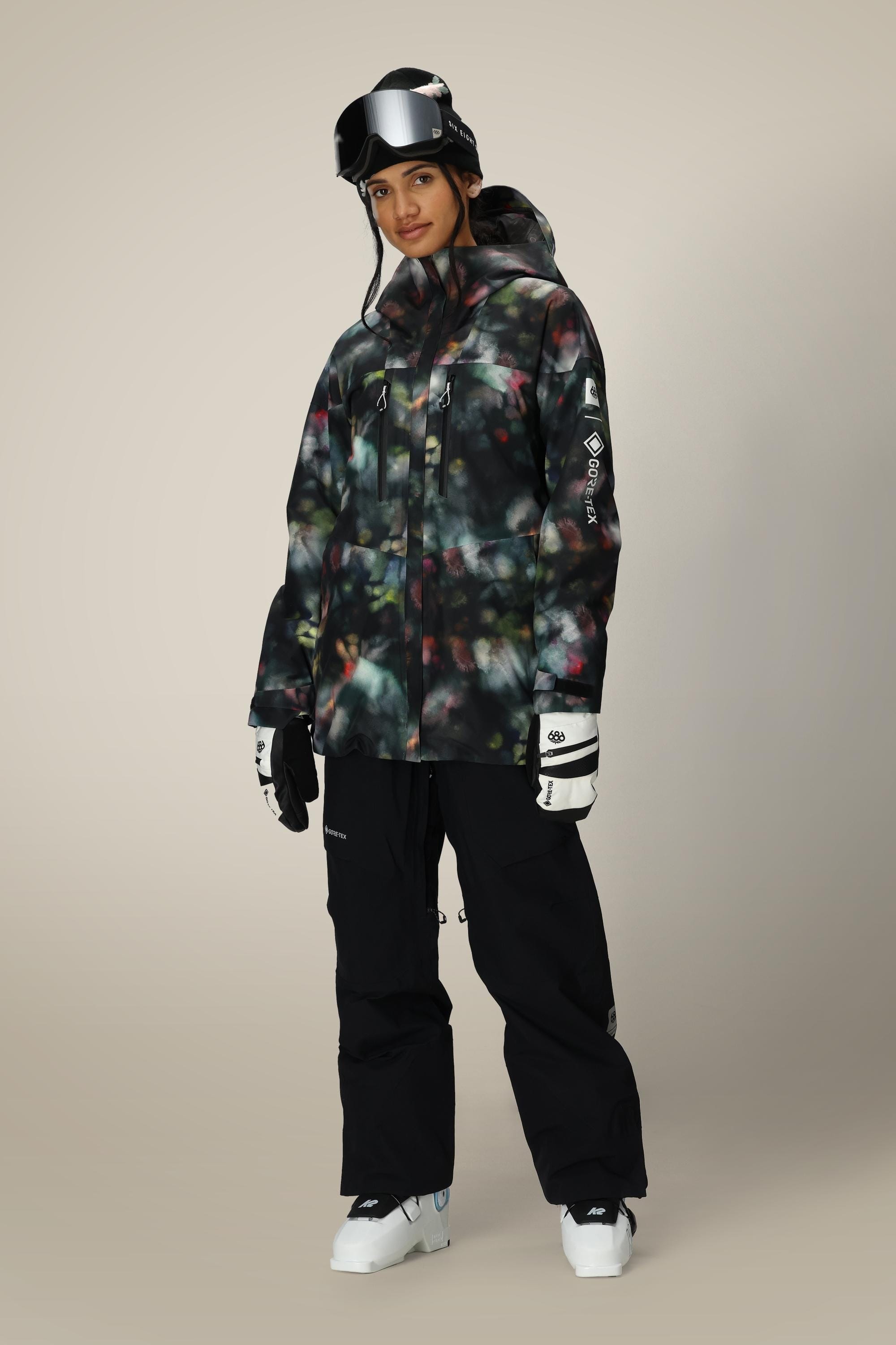 A person wearing the 686 Women's GORE-TEX Skyline Thermagraph® Jacket stands facing forward, paired with black snow pants, white ski gloves, black ski goggles, and white ski boots against a plain background.