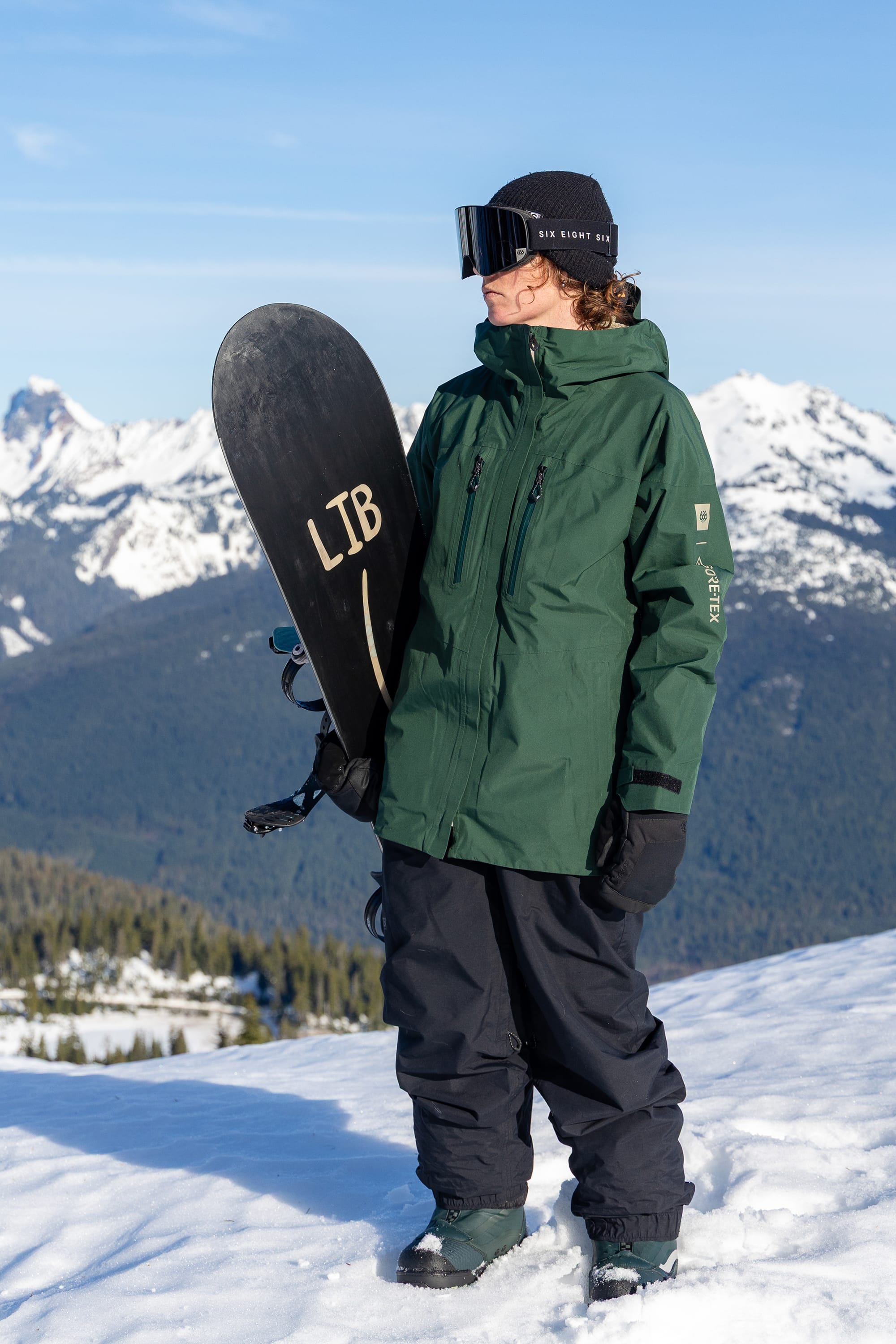 Wearing the 686 Women’s GORE-TEX® Skyline Thermagraph® Jacket, a person stands on a snowy mountain in black snow pants, holding a snowboard upright against a backdrop of snow-covered peaks and trees.