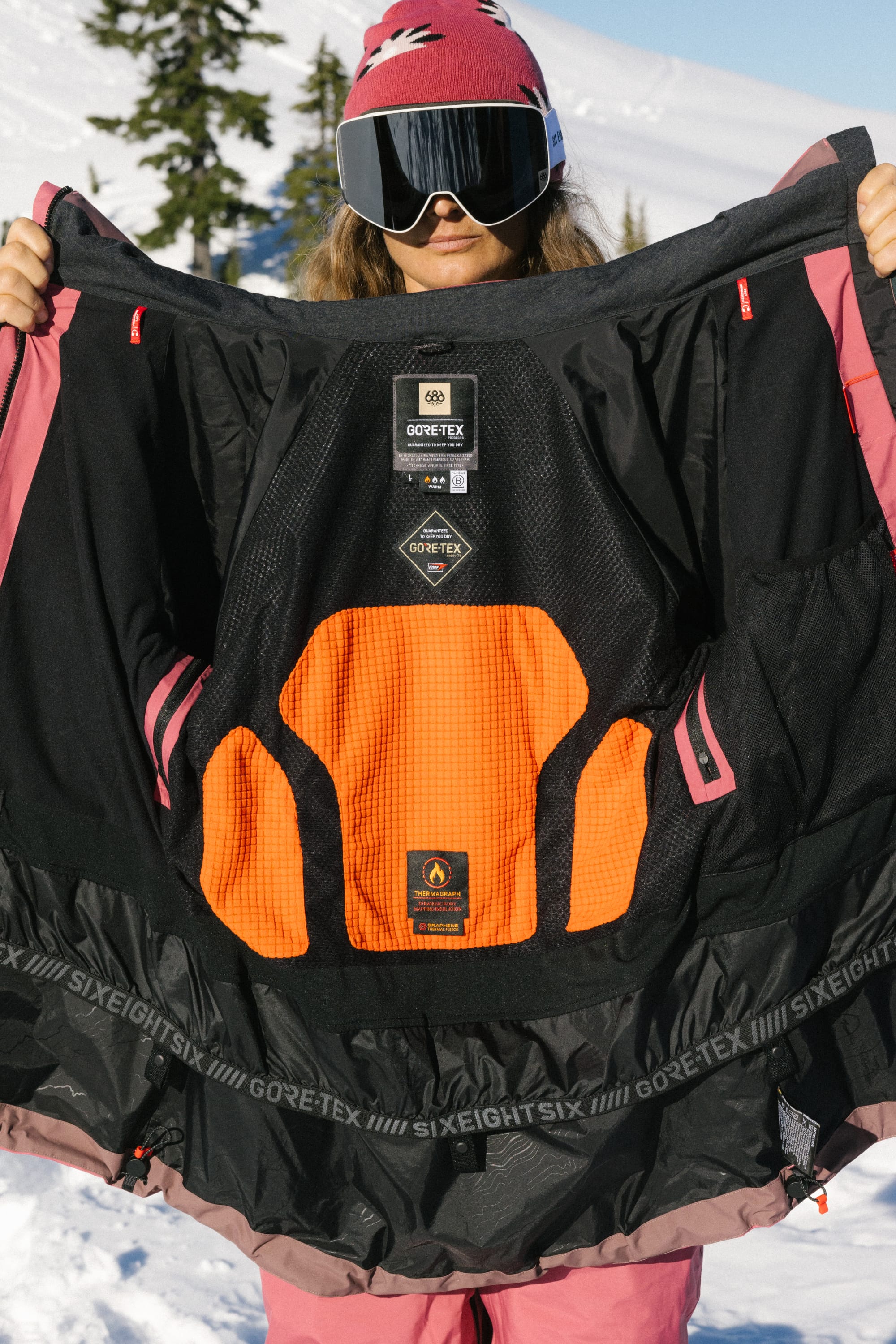 Wearing pink ski gear and goggles, a person displays the detailed lining of the 686 Women's GORE-TEX Skyline Thermagraph® Jacket in black and orange, with snowy mountains and trees in the background.