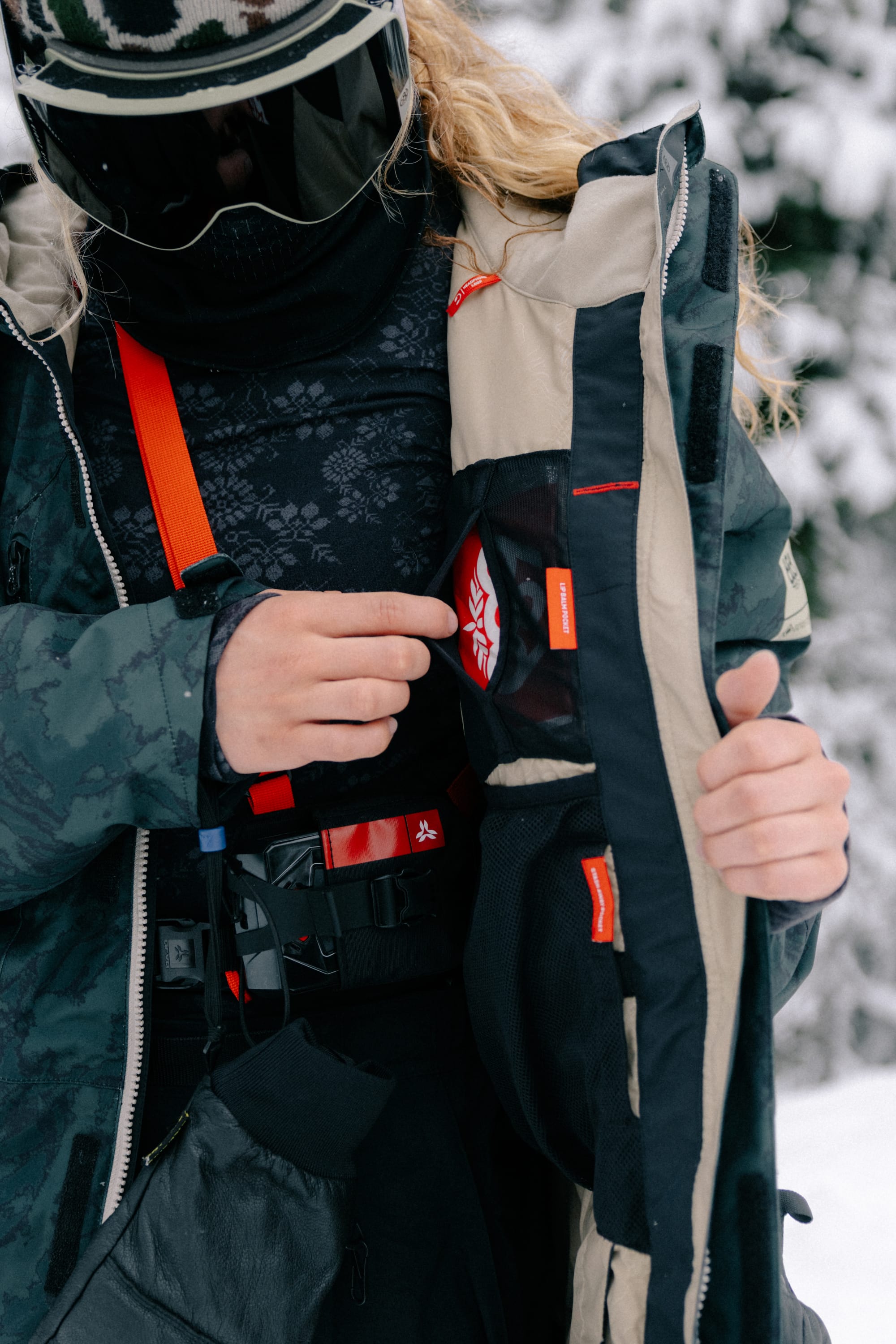 A woman in winter gear opens her 686 Women’s GORE-TEX® Willow™ Insulated Jacket to reveal a pocket with the 686 red and white logo, while snowy trees blur in the background.