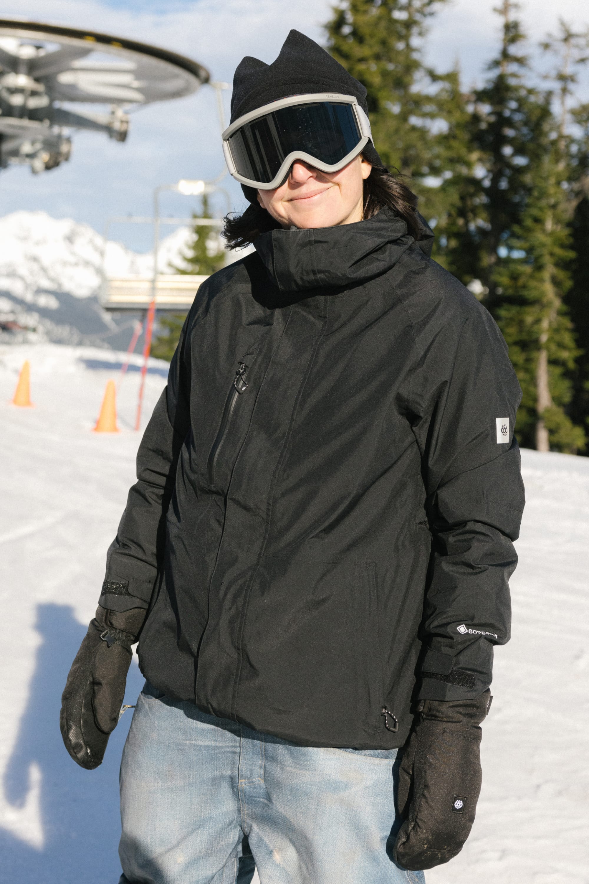A woman wearing the 686 Women’s GORE-TEX® Willow™ Insulated Jacket stands and smiles on a snowy slope, with ski goggles, gloves, a beanie, snow-covered trees, and ski lift equipment visible in the background.