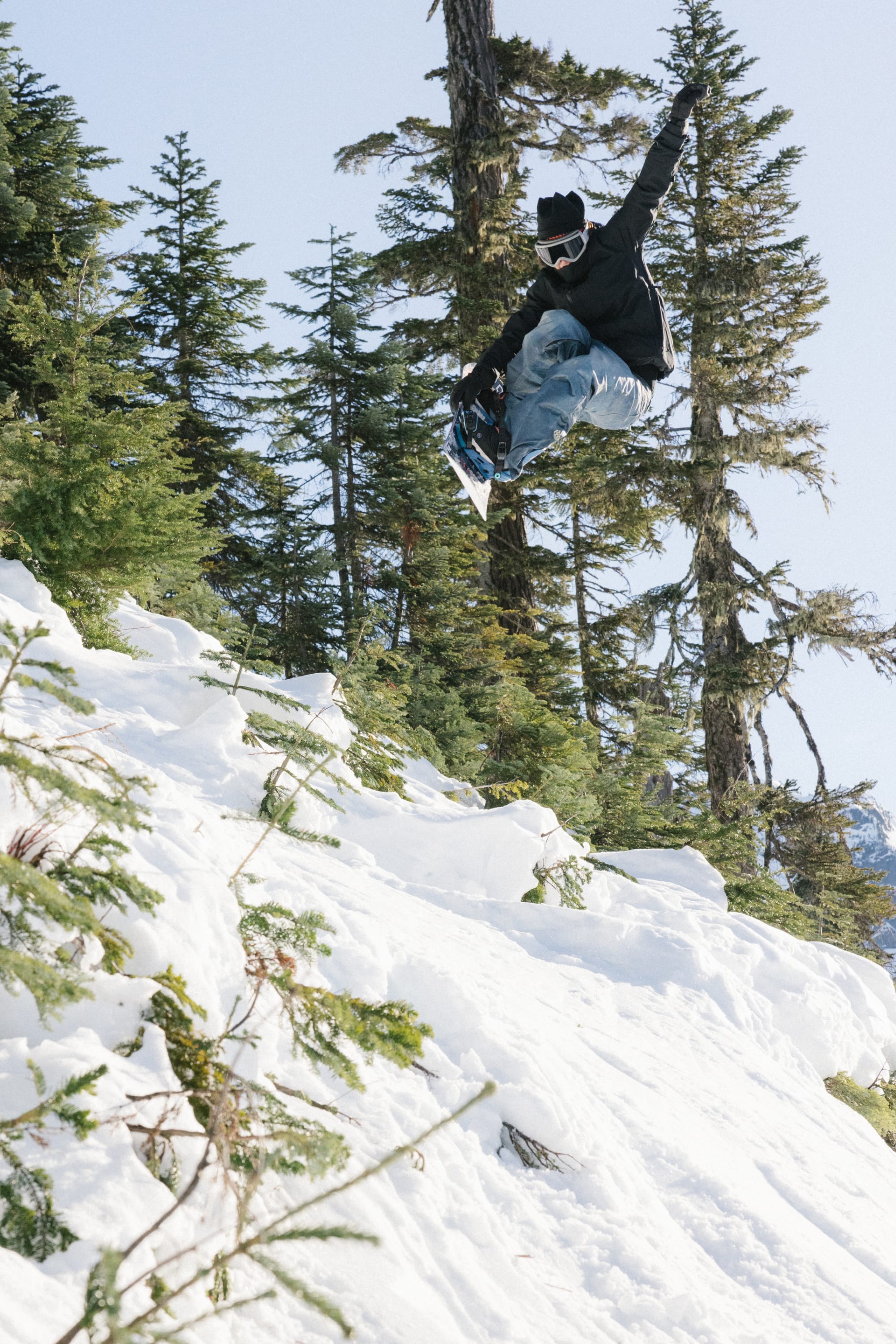 Wearing the 686 Women’s GORE-TEX® Willow™ Insulated Jacket, a snowboarder catches air while performing a jump on a snowy slope, surrounded by evergreen trees beneath a clear sky.