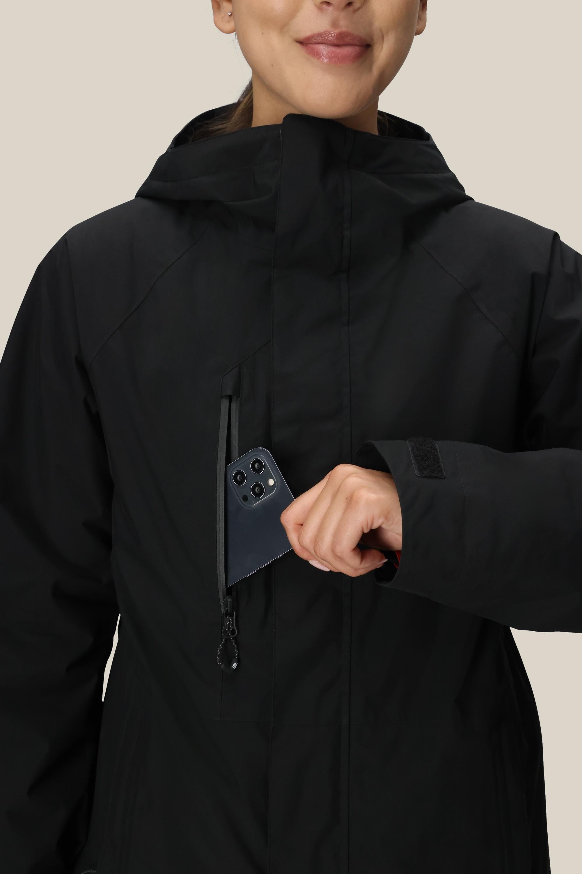 A woman wearing the 686 Women's GORE-TEX Willow™ Insulated Jacket smiles as she places her smartphone into the jacket’s zippered chest pocket.