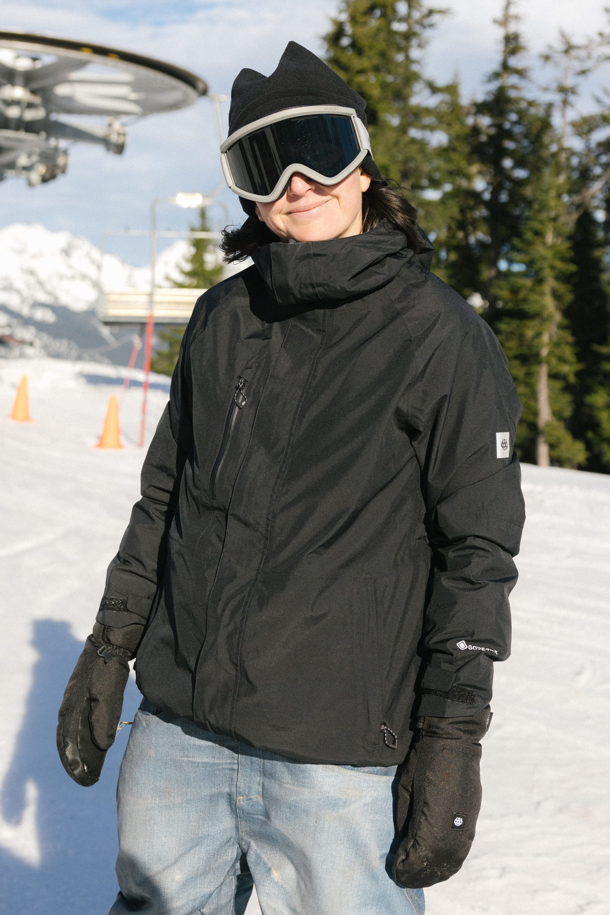 Wearing a 686 Women's GORE-TEX Willow™ Insulated Jacket, ski gloves, goggles, and a beanie, a person stands on a snowy slope with trees and ski lift equipment in the background.