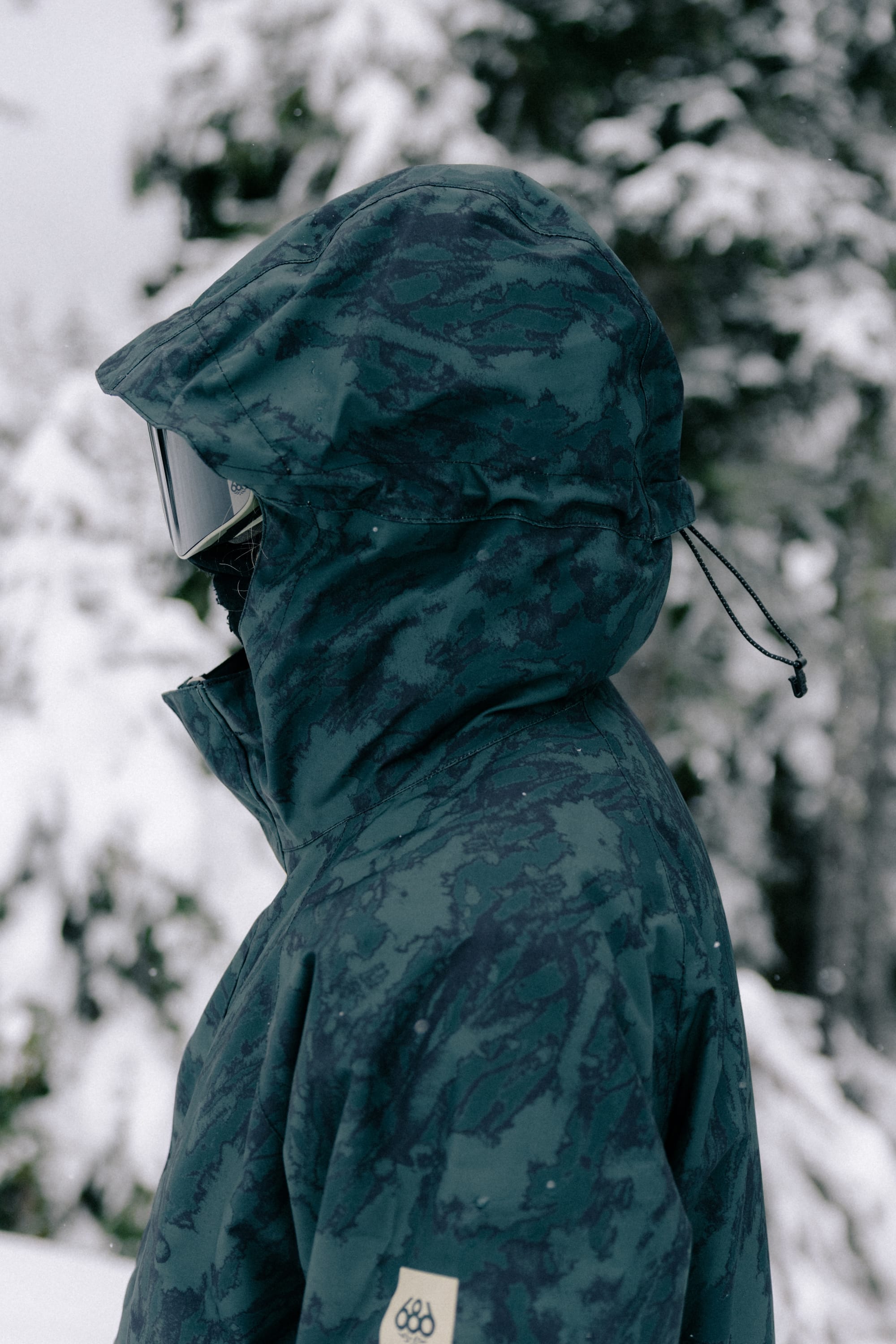 Wearing the 686 Women’s GORE-TEX® Willow™ Insulated Jacket with the hood up and ski goggles, a person stands in a snowy outdoor setting surrounded by trees.