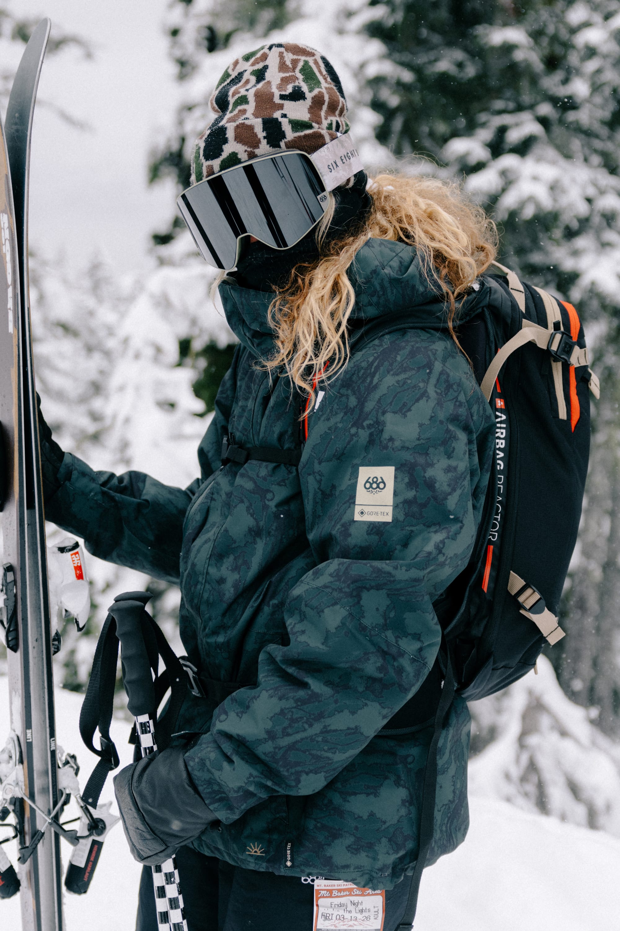 Wearing the 686 Women’s GORE-TEX® Willow™ Insulated Jacket, a person in winter gear—with a patterned beanie, reflective ski goggles, and backpack—holds skis and poles amidst a snowy forest backdrop.