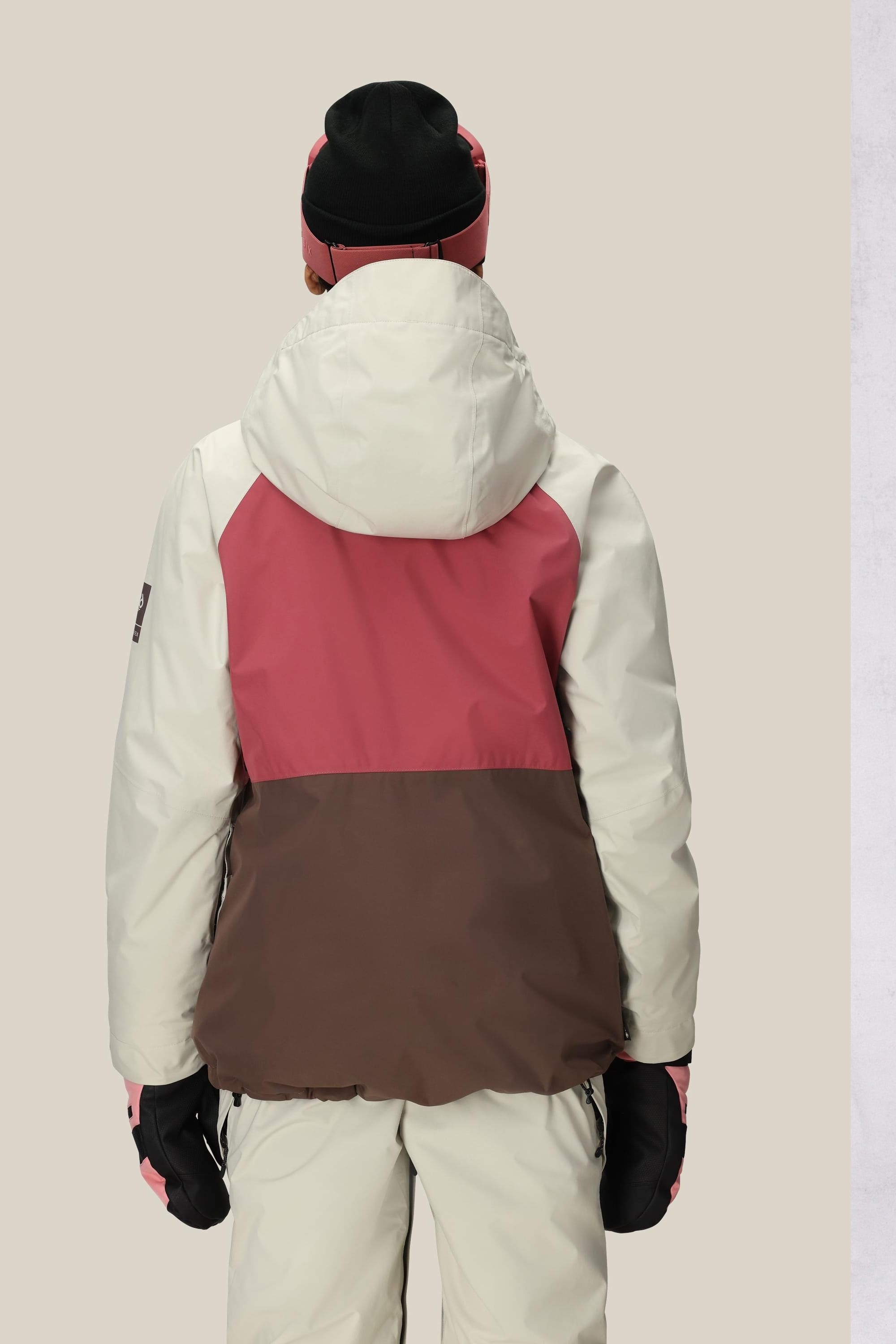 A person is seen from the back wearing the 686 Women's GORE-TEX Willow™ Insulated Jacket in cream, pink, and brown by 686, paired with cream pants, black gloves, and a black beanie against a plain beige background.