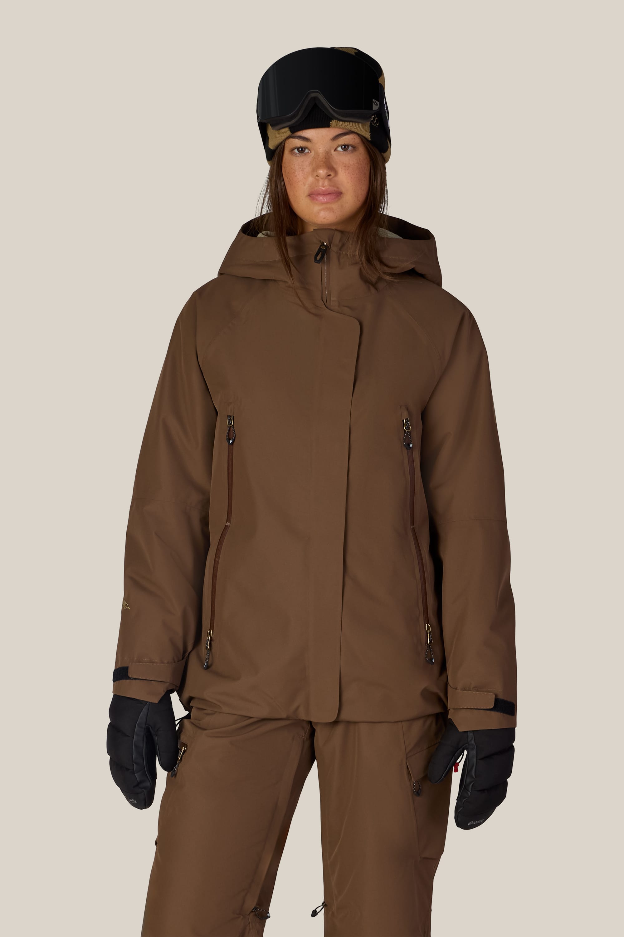 A person is wearing the 686 Women’s Whisper™ Insulated Jacket by 686, along with matching pants, black gloves, and ski goggles while standing facing forward against a plain light background.