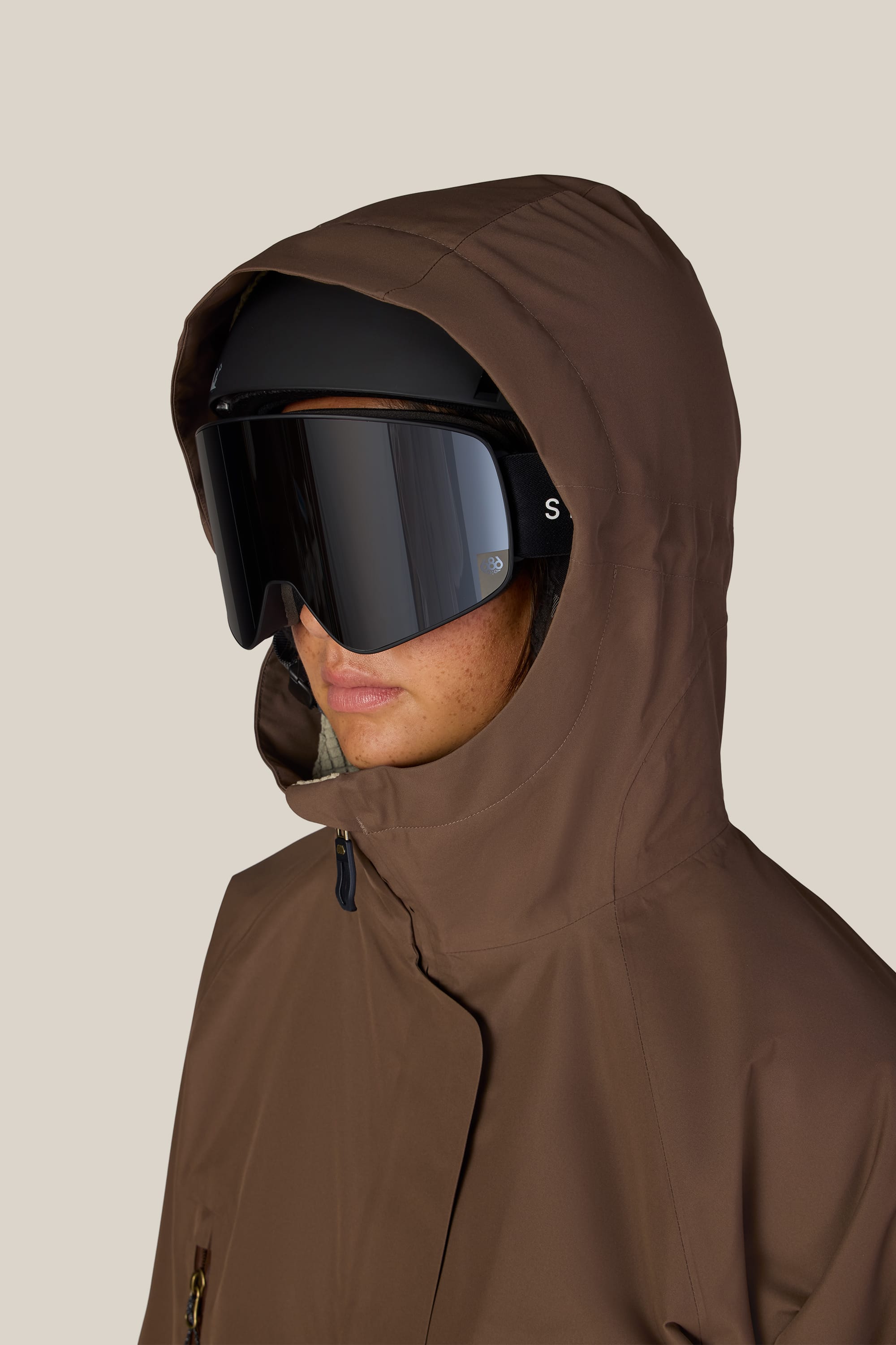 A person is seen facing left, wearing the 686 Women’s Whisper™ Insulated Jacket in brown with a hood up and black ski goggles. The background is plain and light-colored, and only the lower part of the face is visible.