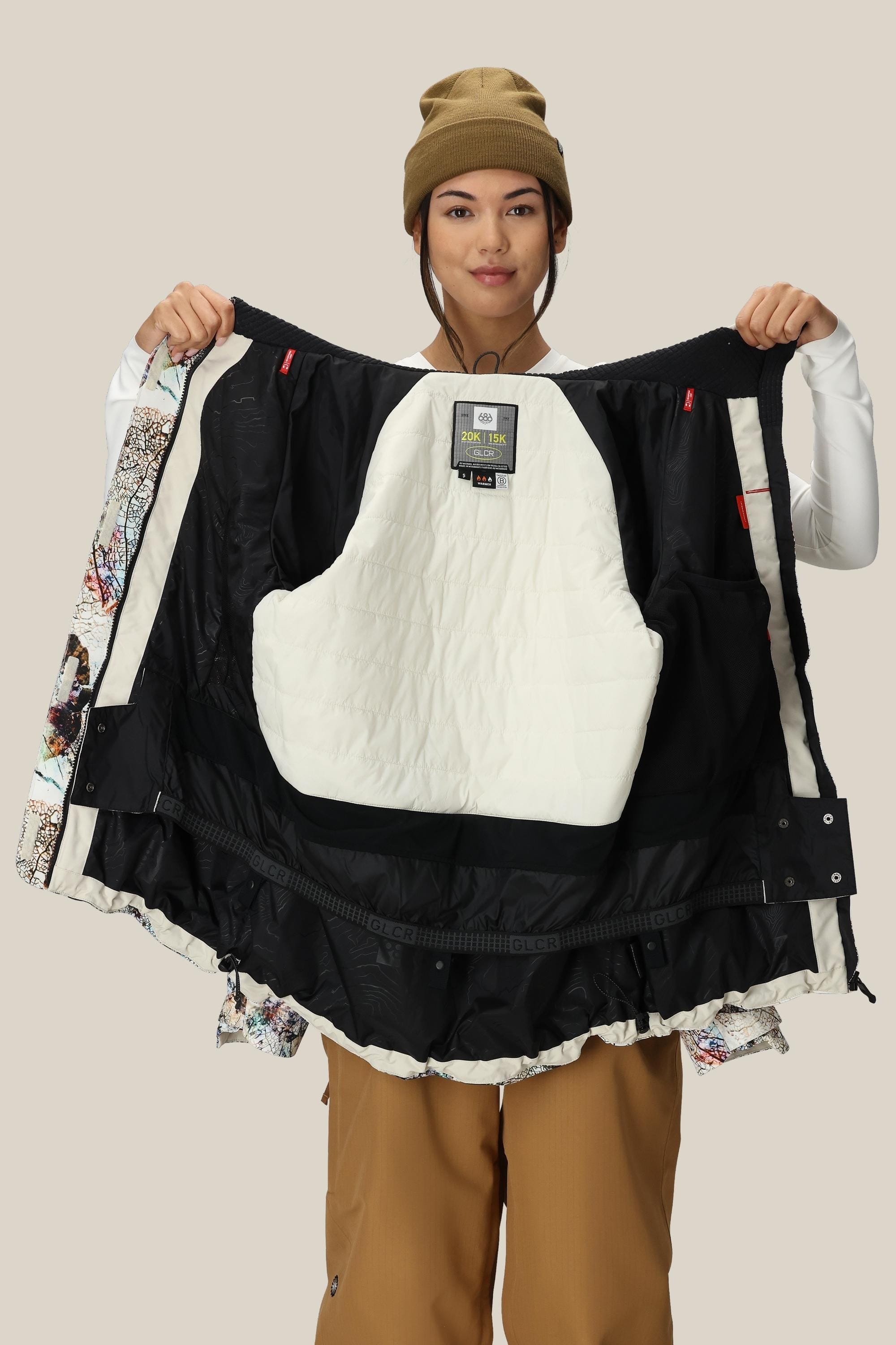 A woman in a tan beanie and brown pants, facing the camera and smiling, holds open the 686 Women's Whisper™ Insulated Jacket to display its white and black quilted interior lining.
