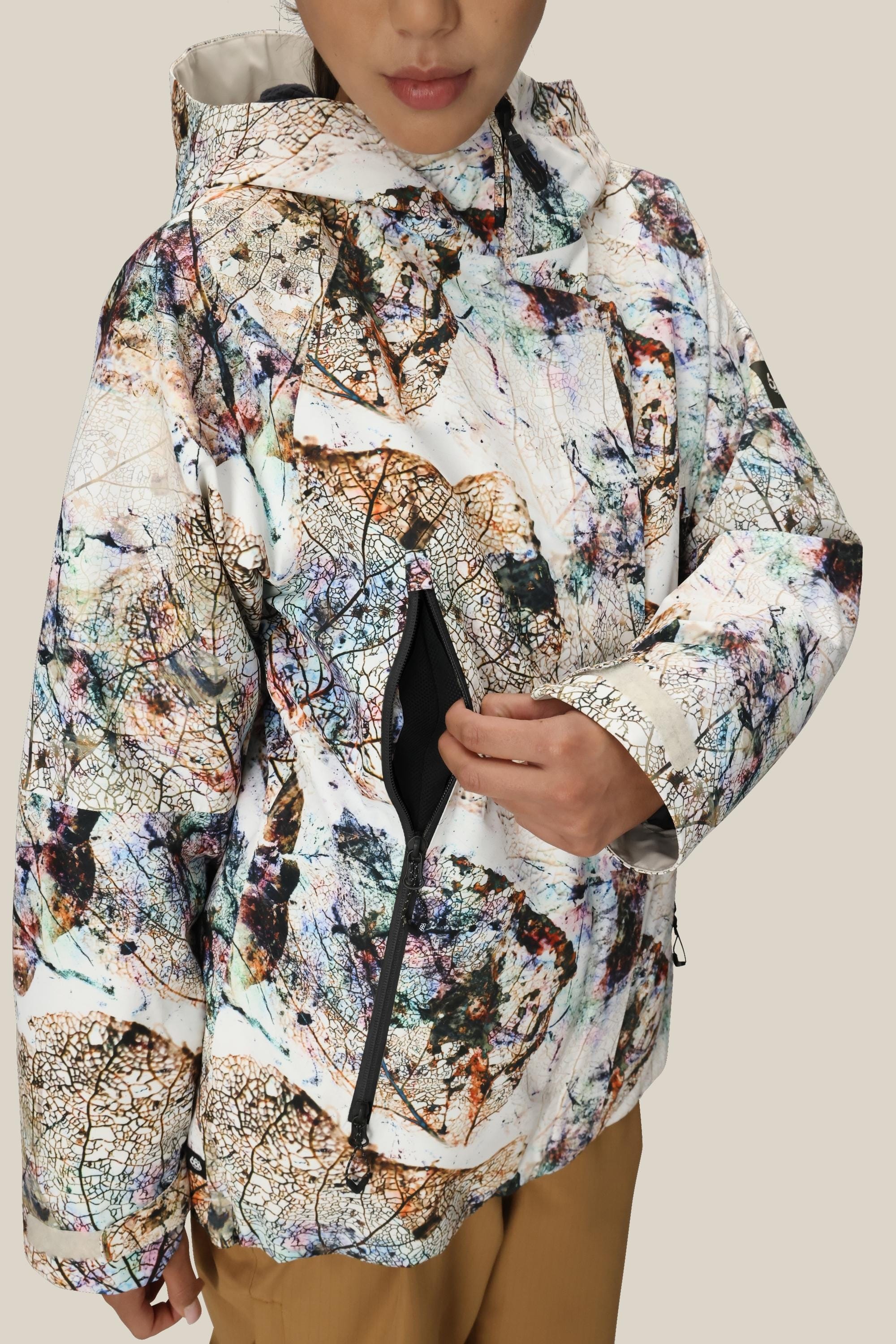 A person in a 686 Women's Whisper™ Insulated Jacket with an abstract pattern and hood unzips a side pocket. Only the lower half of their face is visible against a plain beige background.