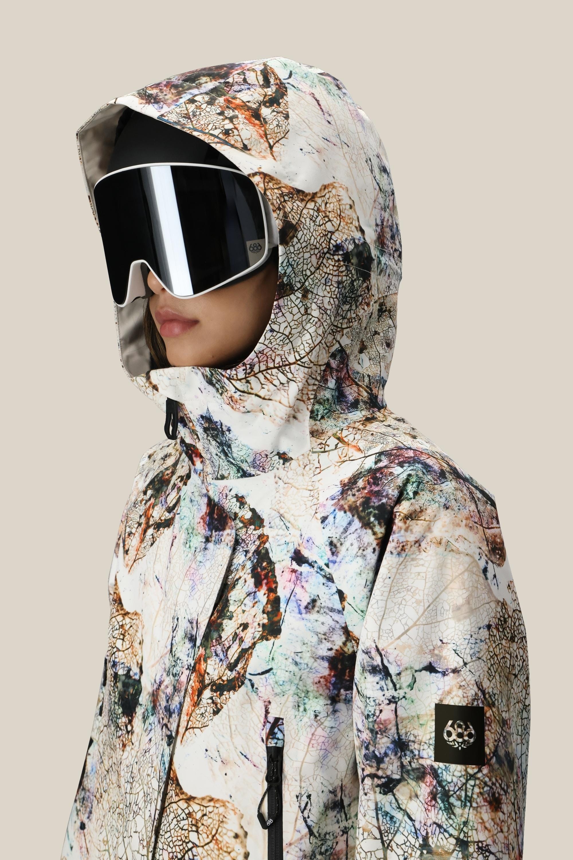 A person wearing the 686 Women's Whisper™ Insulated Jacket—an abstract-patterned, polyester waterproof coat with a hood and 686 logo patch—pairs it with large black ski goggles, standing against a plain beige background.