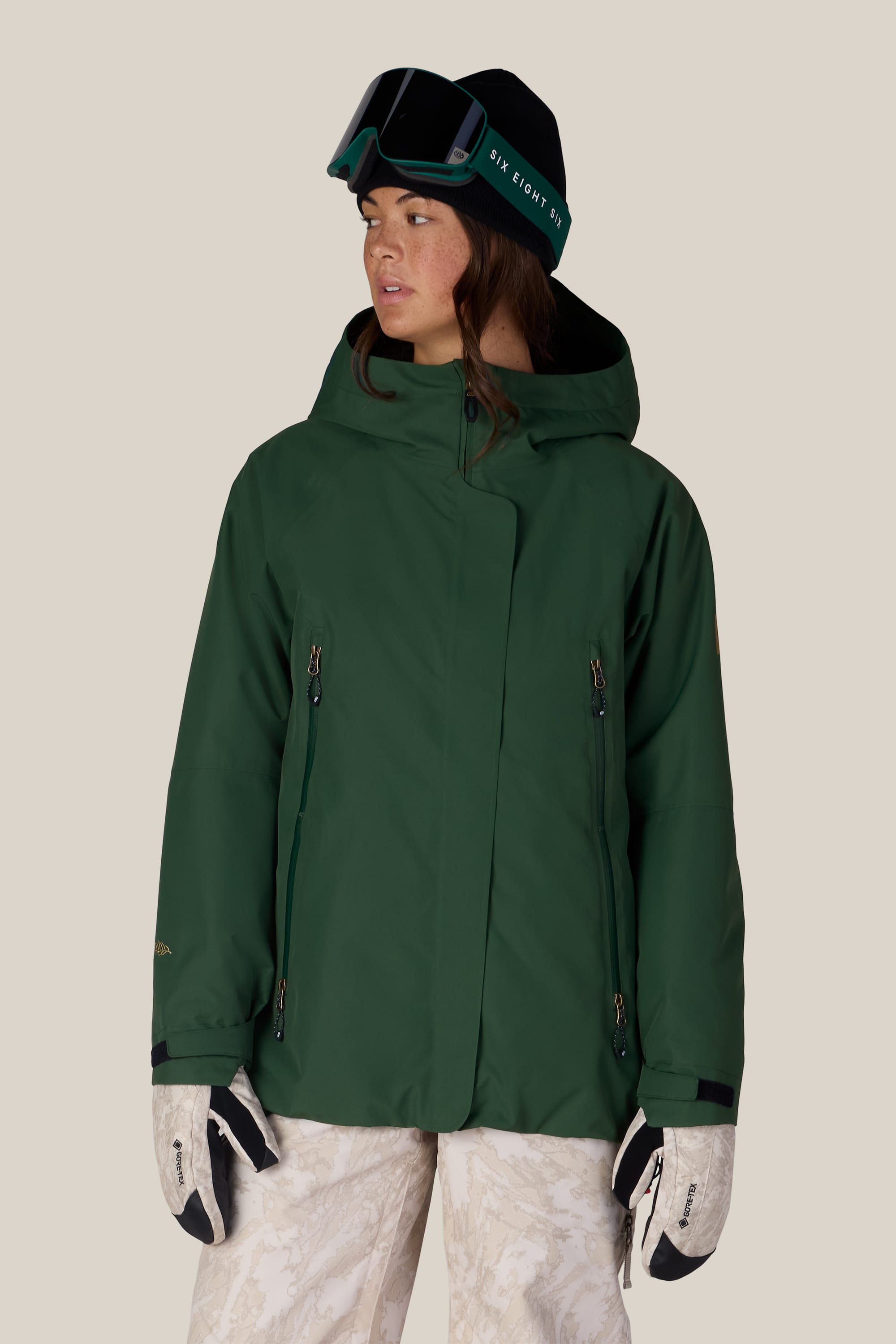 Wearing the 686 Women’s Whisper™ Insulated Jacket in green, along with beige patterned snow pants, white gloves, a black beanie, and ski goggles, a person stands against a plain beige background.