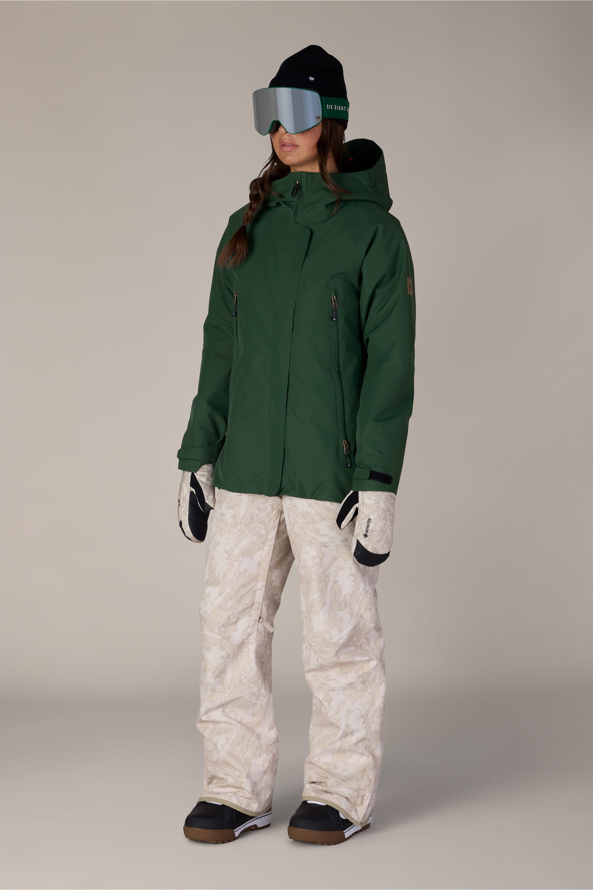 A person wearing the 686 Women’s Whisper™ Insulated Jacket by 686, patterned white snow pants, black boots, gloves, large ski goggles, and a black beanie stands against a plain background.