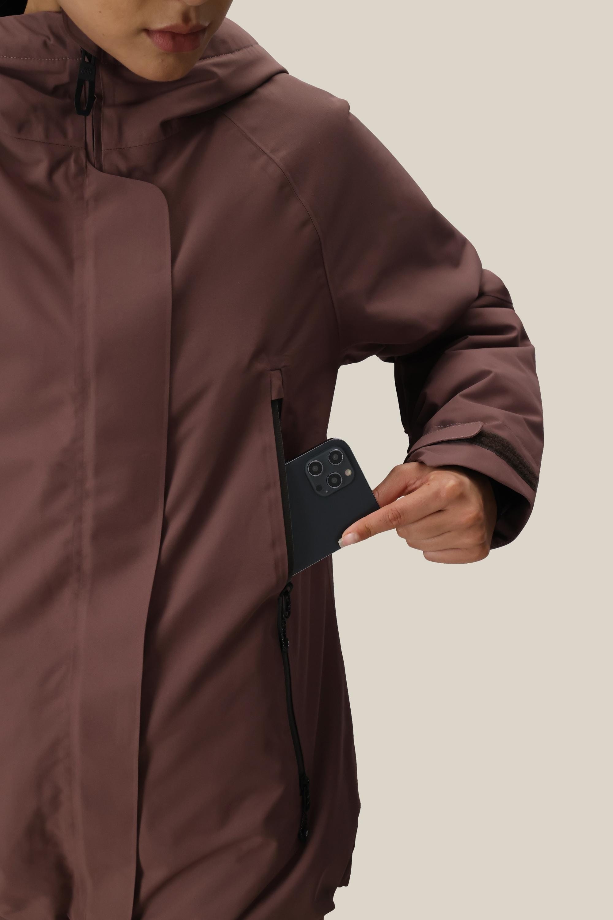 A woman in a 686 Women's Whisper™ Insulated Jacket slips a black smartphone into the zippered chest pocket, set against a plain, light background.