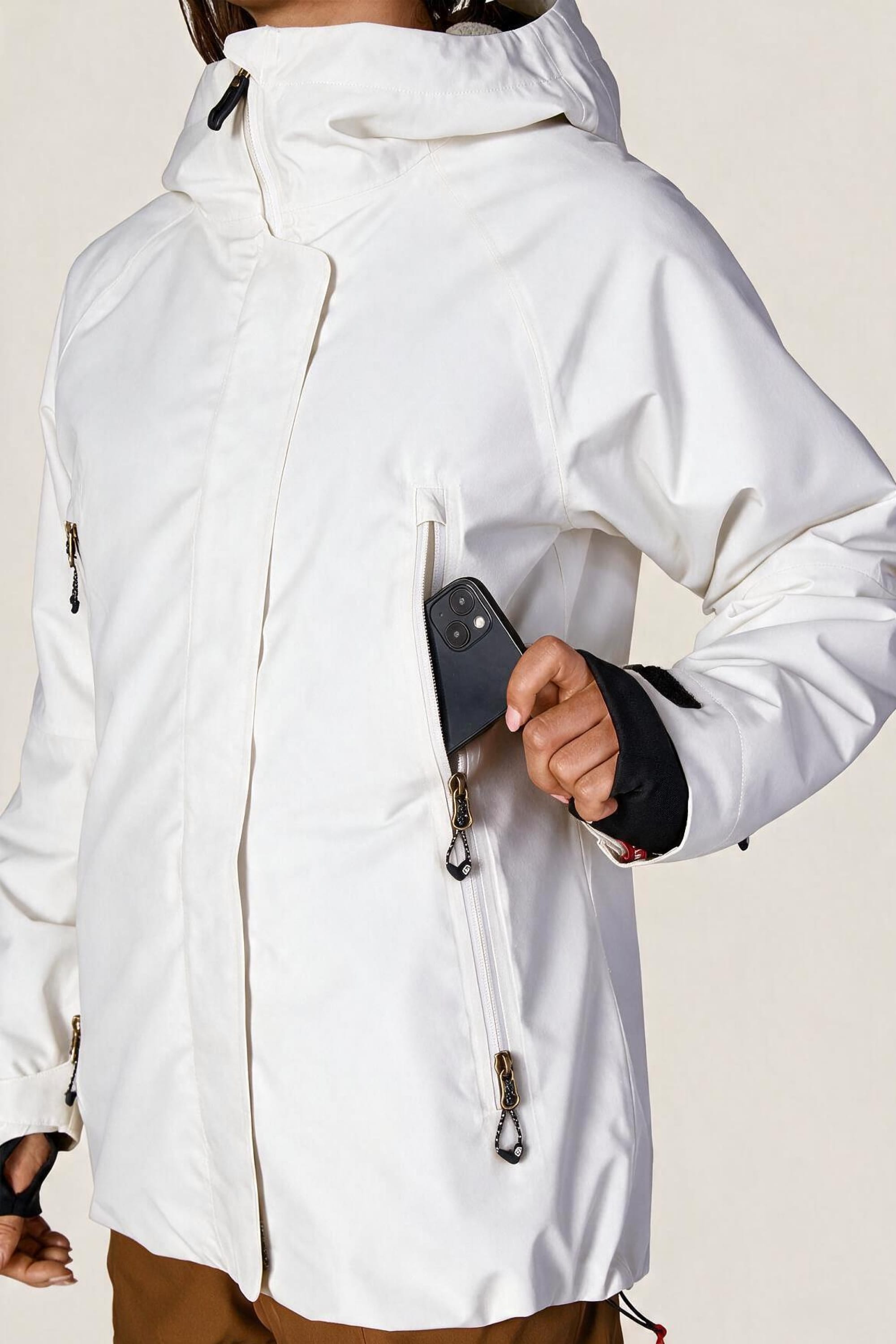 A person wearing the 686 Women’s Whisper™ Insulated Jacket in white and brown pants puts a black smartphone into the jacket’s zippered chest pocket.