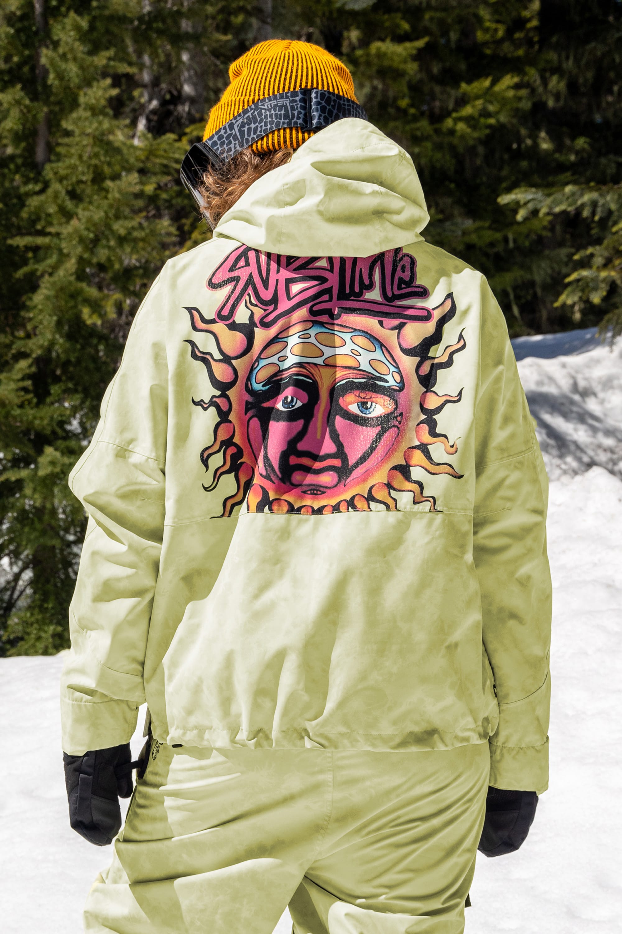 A person stands outside in the snow among trees, wearing the 686 Womens Sublime Shell Anorak—a yellow-green waterproof jacket with a bold sun and face graphic and “Sublime” text from the brand 686.
