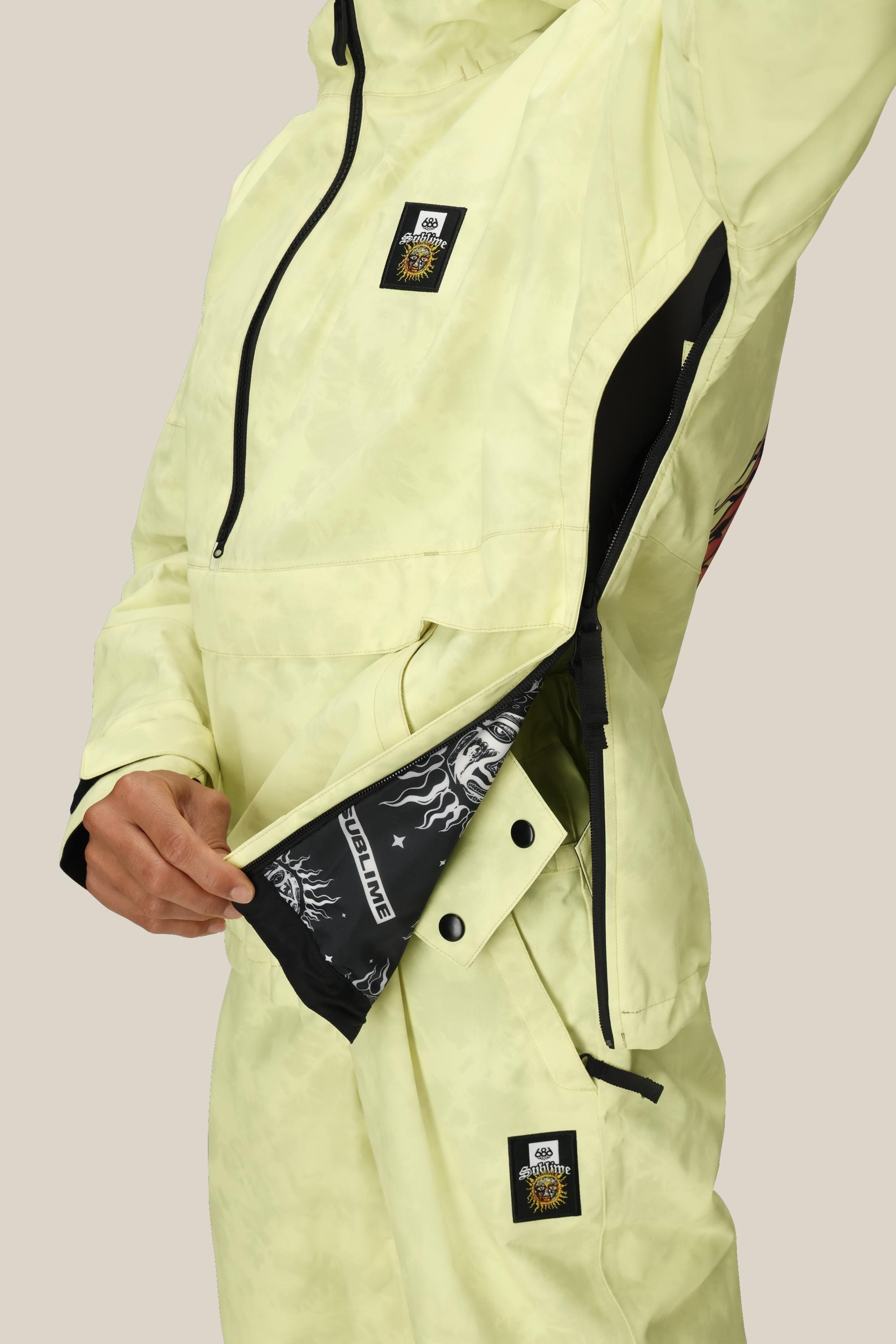 A person wearing the 686 Womens Sublime Shell Anorak unzips a large side zipper, revealing a black inner lining with white graphic designs. The pale yellow jacket and pants feature square black-and-white patches on the chest and pants.