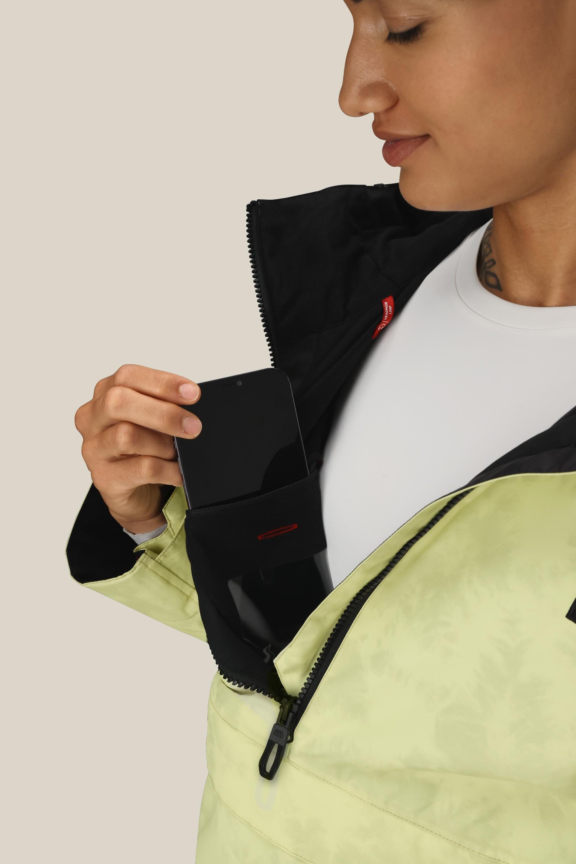 Wearing the 686 Womens Sublime Shell Anorak in light green, a woman partially unzips the jacket to place a black smartphone into its inner chest pocket. The background is plain and light-colored.
