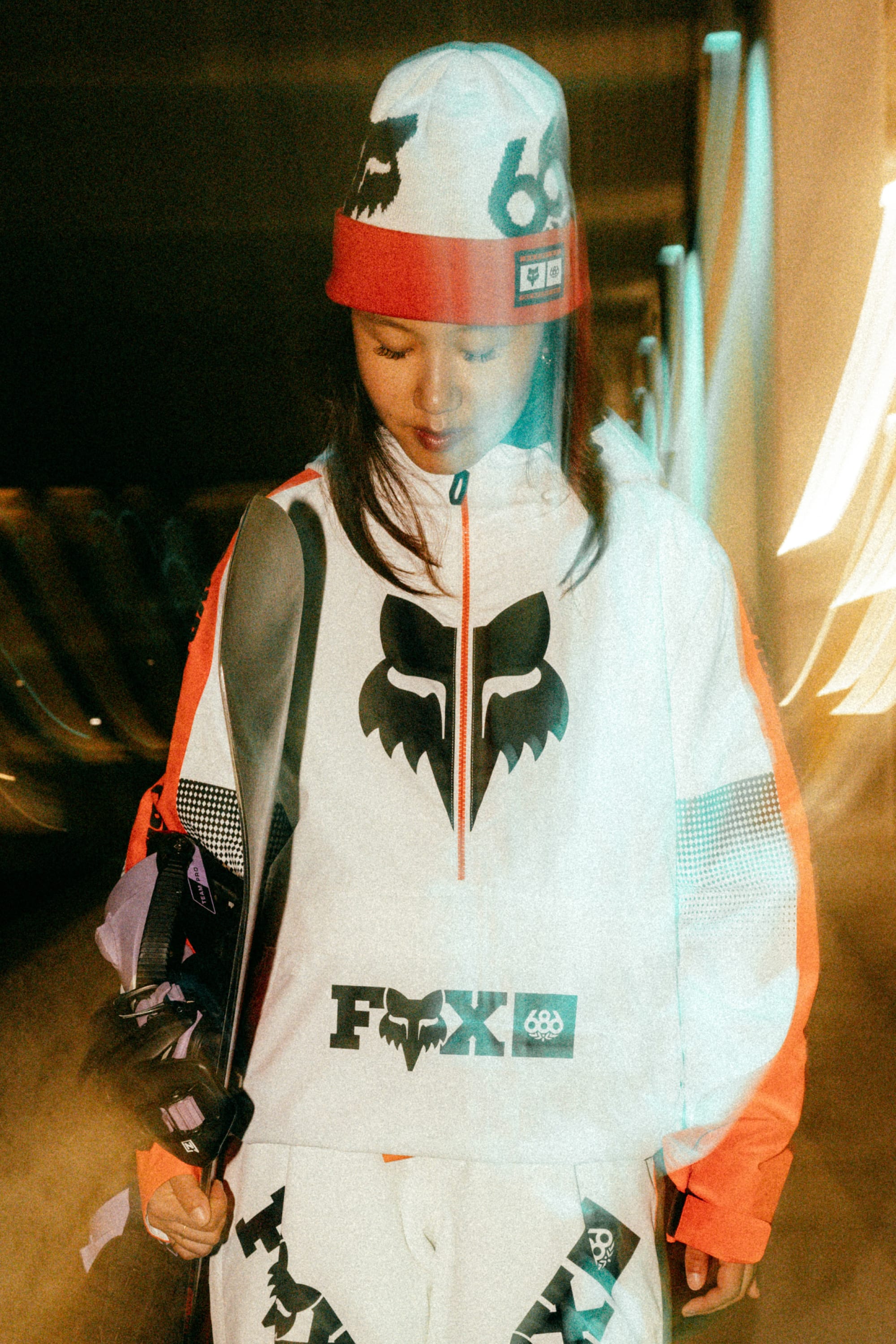 A woman in the 686 Women's Fox Racing Shell Anorak and a 686 beanie stands holding a snowboard in a dimly lit space with blurred lights.