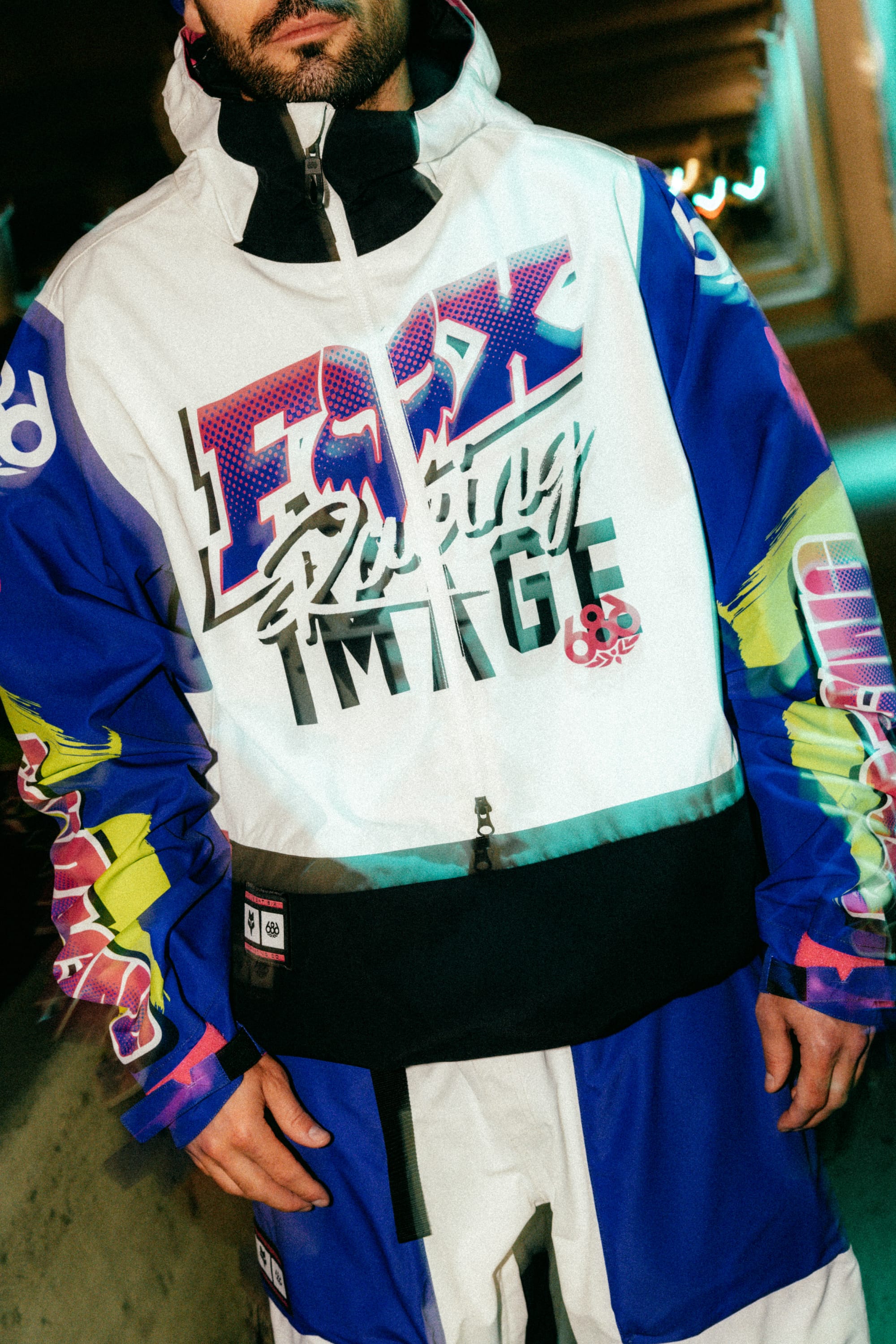 A person wearing the 686 Men's Fox Racing Shell Anorak, featuring bold graphics and FOX Racing branding, stands outdoors at night under artificial lights with their face partially visible.