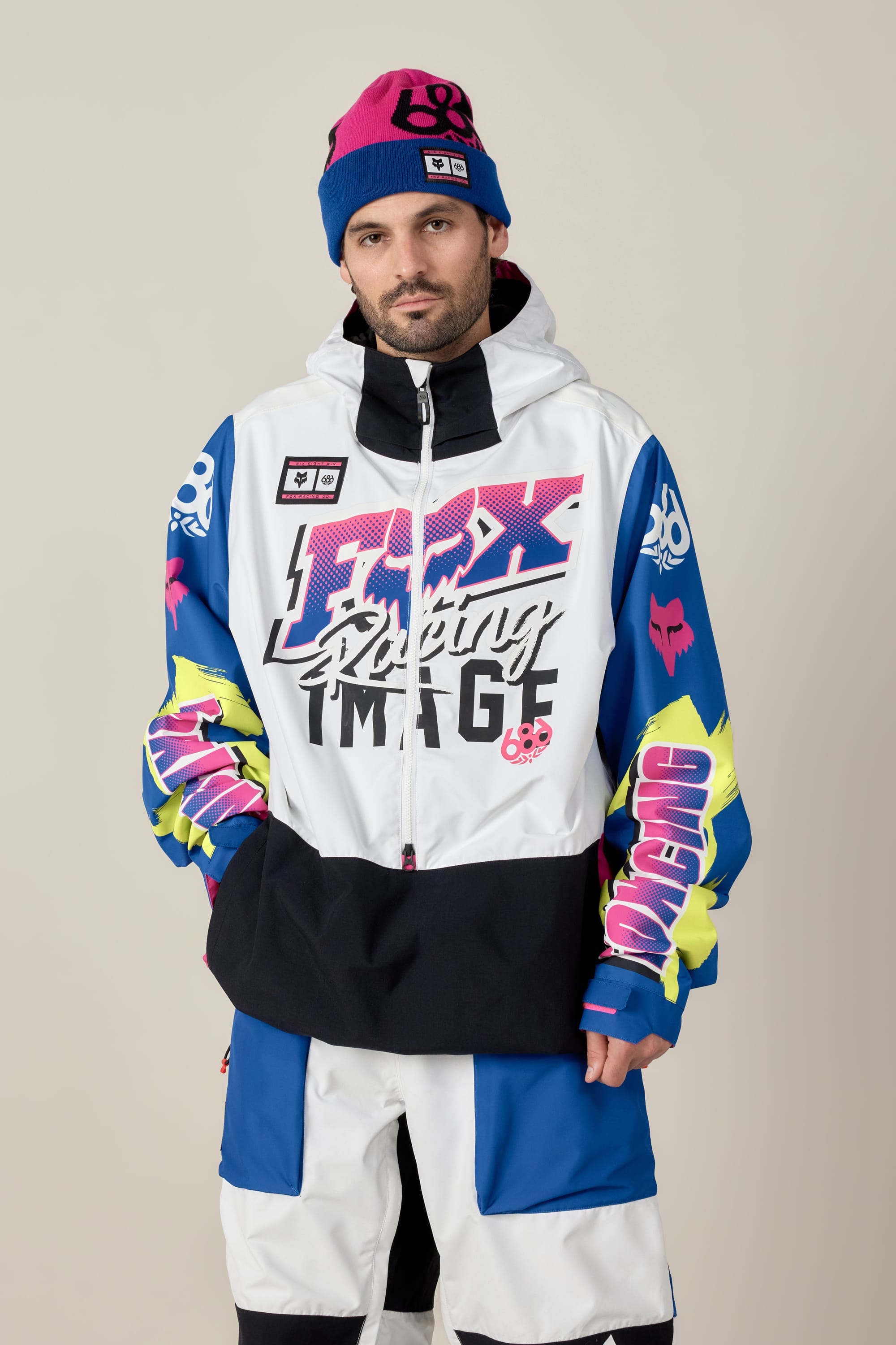 A man models the 686 Men's Fox Racing Shell Anorak by 686, a white, blue, and neon racing-inspired jacket with bold graphics, paired with a blue-pink beanie, standing against a plain light background.