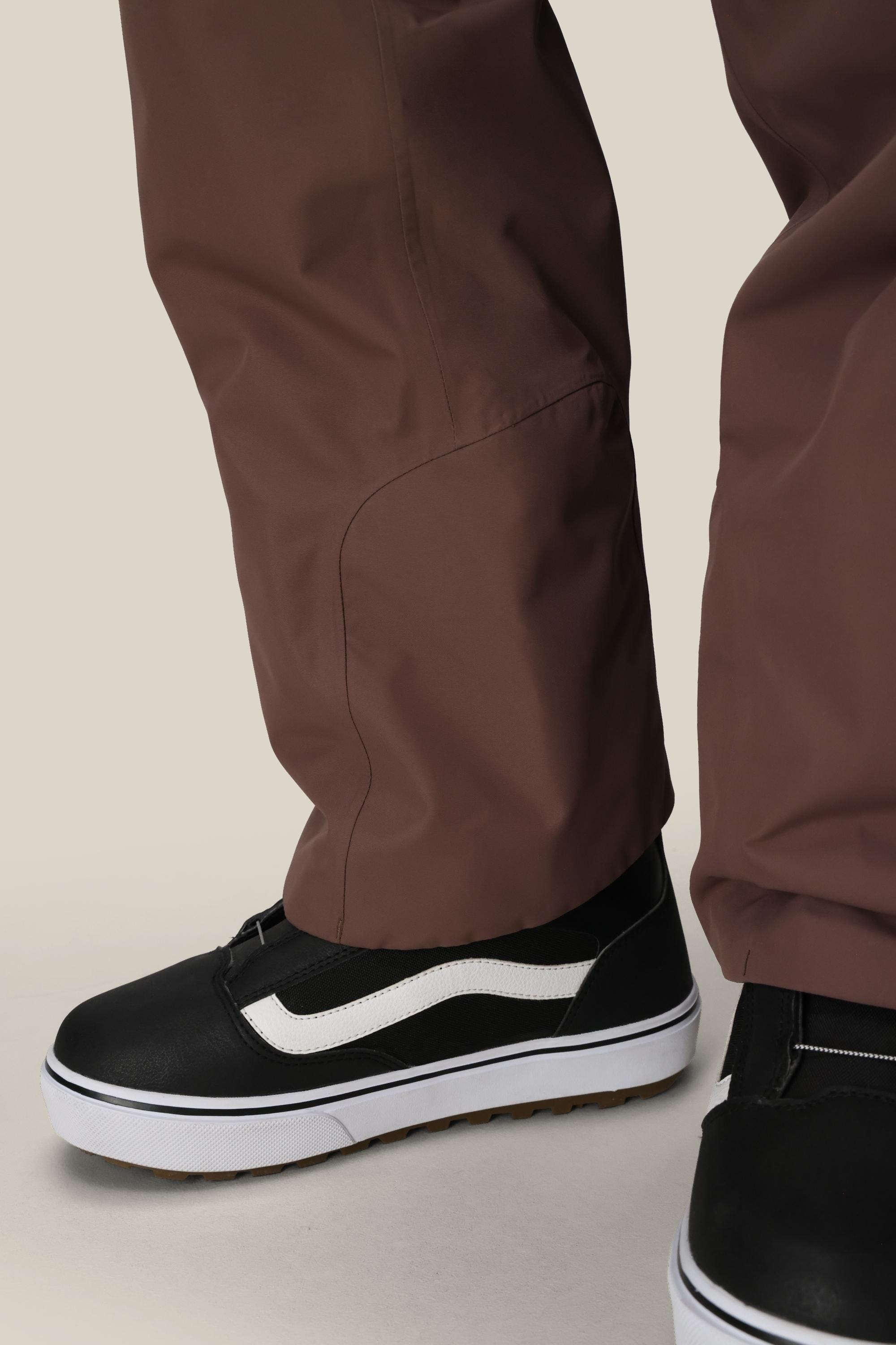 A close-up of a person wearing 686 Women's Geode Thermagraph® Pant by 686, paired with black slip-on sneakers featuring white soles and a stripe detail, standing on a neutral background.