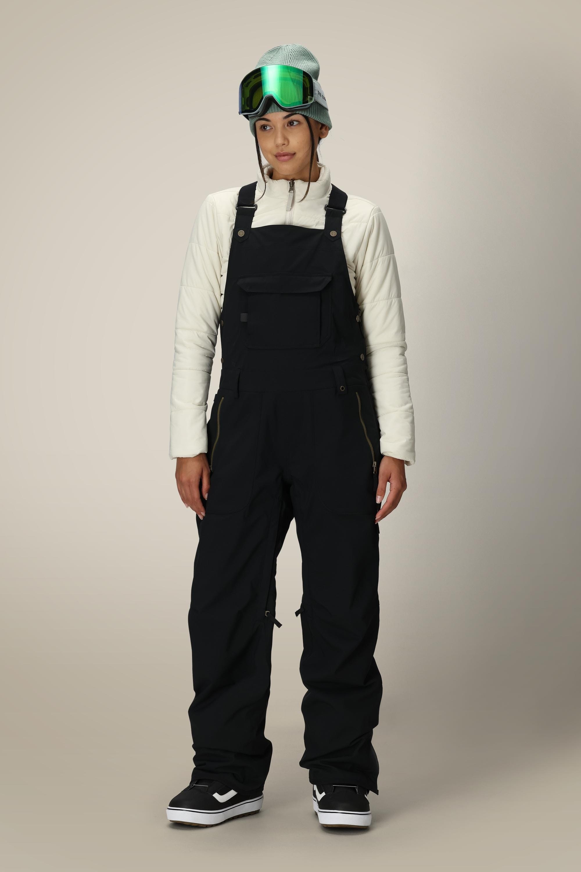 A person wearing a white jacket, the 686 Women’s Black Magic™ Insulated Bib by 686, a teal beanie, and green ski goggles stands facing forward against a plain background.