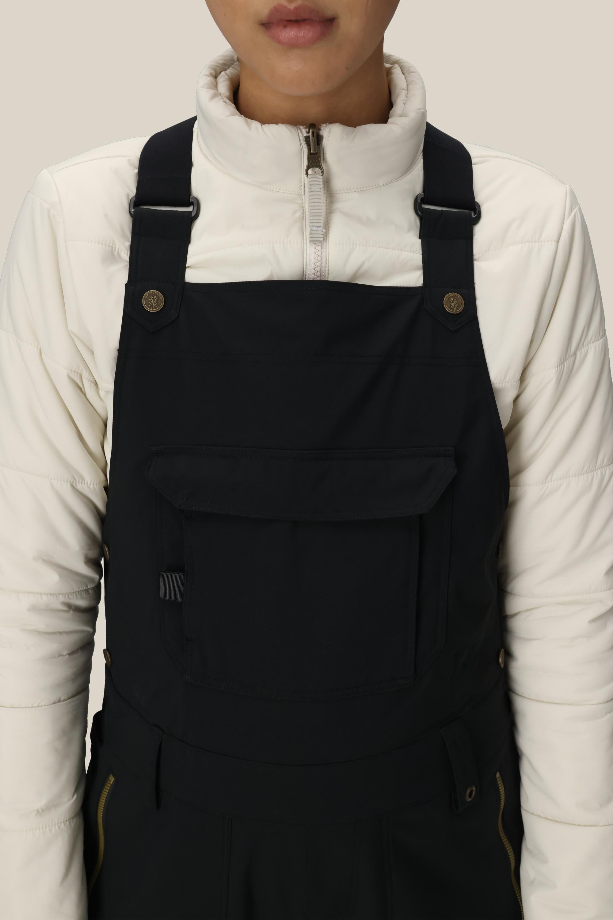 A person is wearing a white zip-up jacket and the 686 Women's Black Magic™ Insulated Bib by 686, shown from shoulders to waist against a plain beige background.
