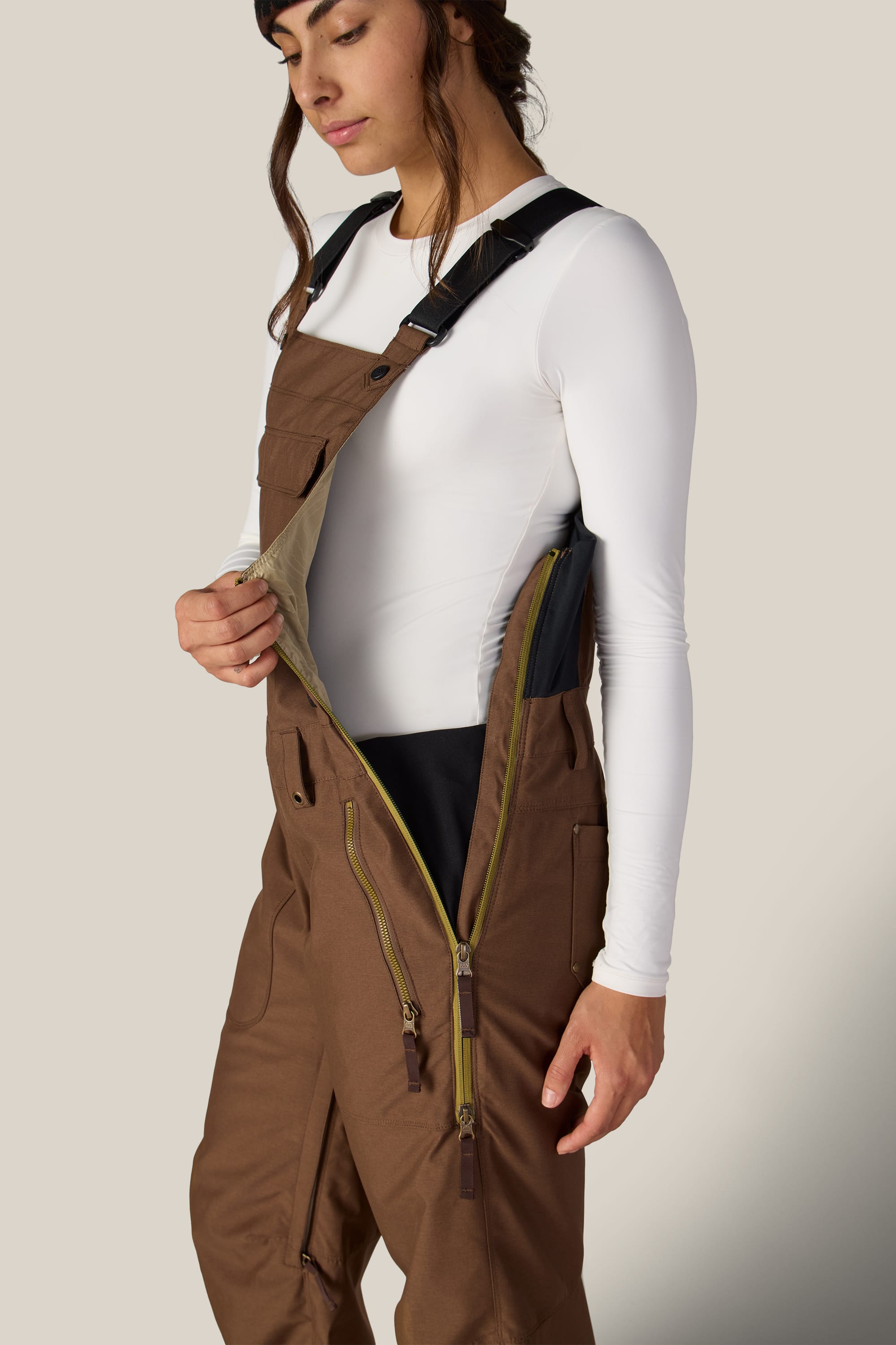 A person wearing the 686 Women’s Black Magic™ Insulated Bib by 686 with one strap unfastened, revealing a white long-sleeve shirt underneath. They are unzipping the side of the bib to show the inner lining.