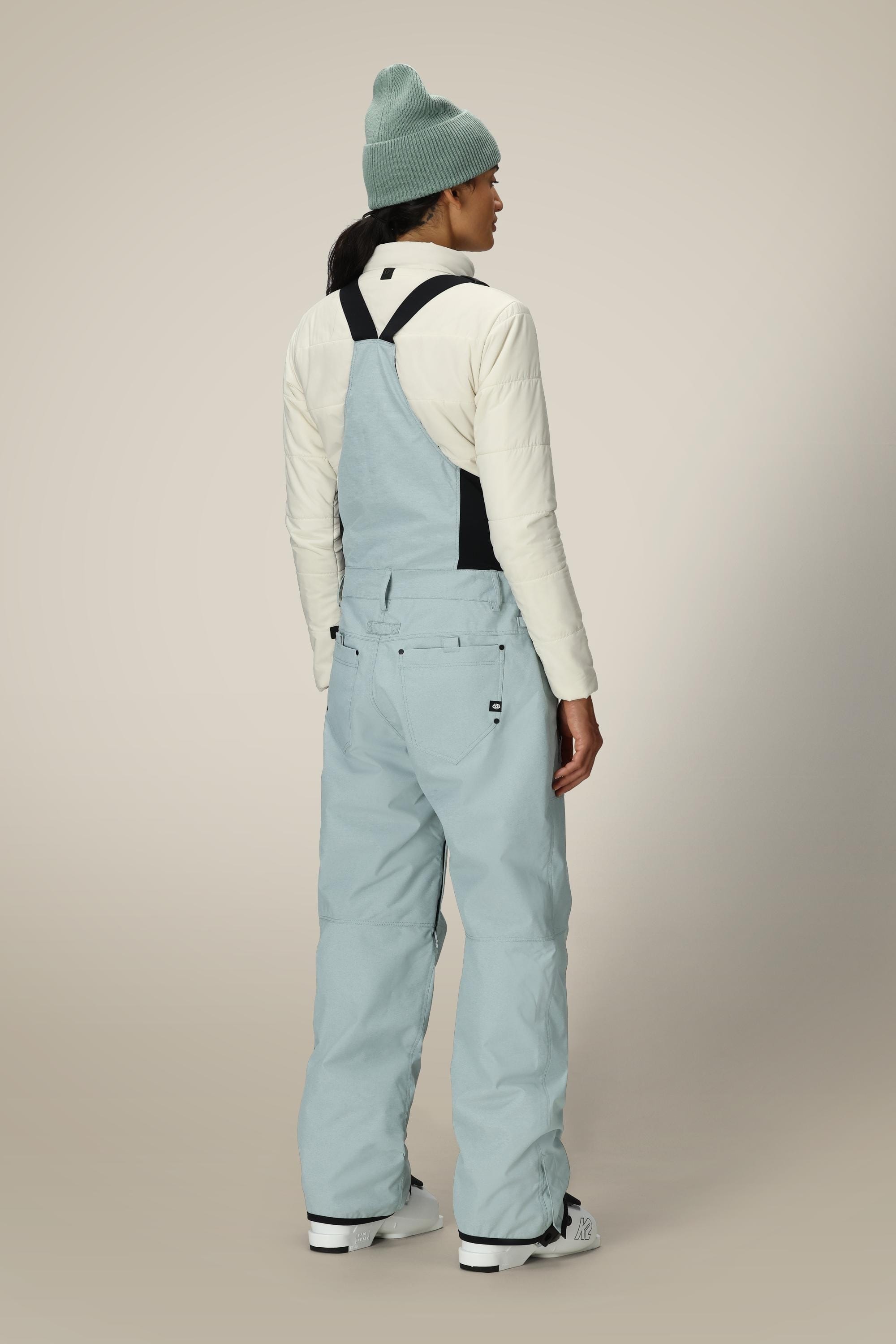 A person stands facing away, wearing a light blue beanie, off-white quilted jacket, white shoes, and 686 Women's Black Magic™ Insulated Bib snow pants with black straps by 686, against a neutral background.