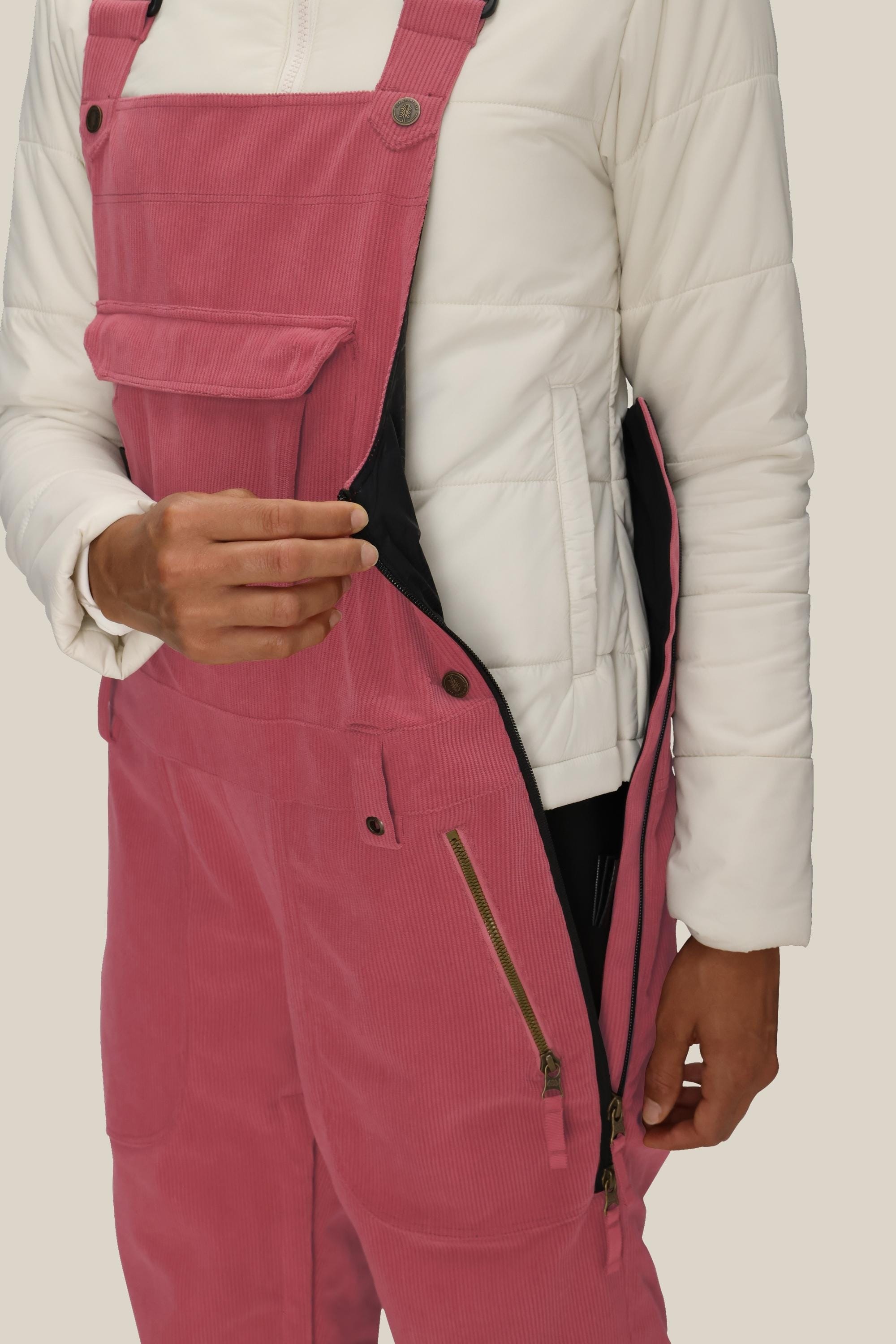 A person wearing a white quilted jacket and 686 Women's Black Magic™ Insulated Bib unzips a side zipper on the bib, revealing its black inner lining. Only the torso and one hand are visible.