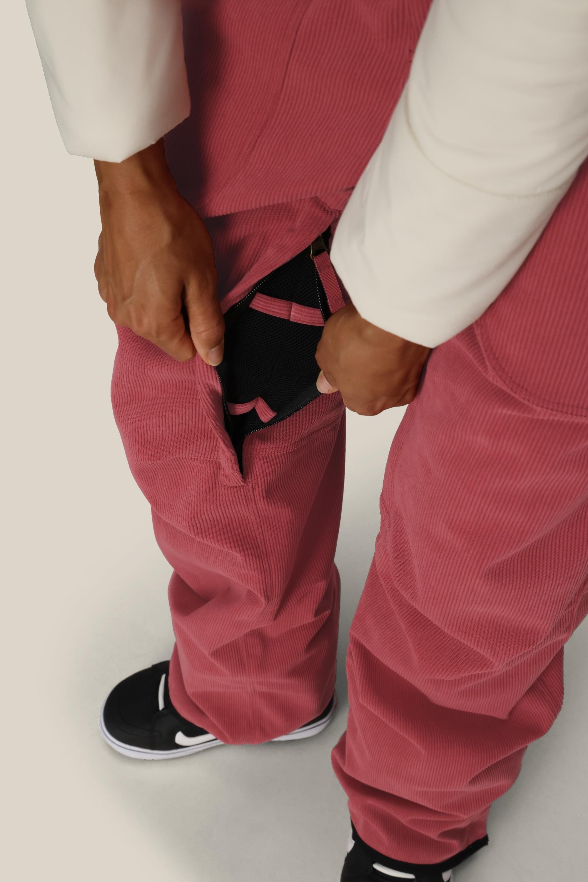 A person in pink corduroy pants and black sneakers opens a hidden zipper pocket, revealing the 686 Women's Black Magic™ Insulated Bib with pink straps inside. The background is plain and light-colored.