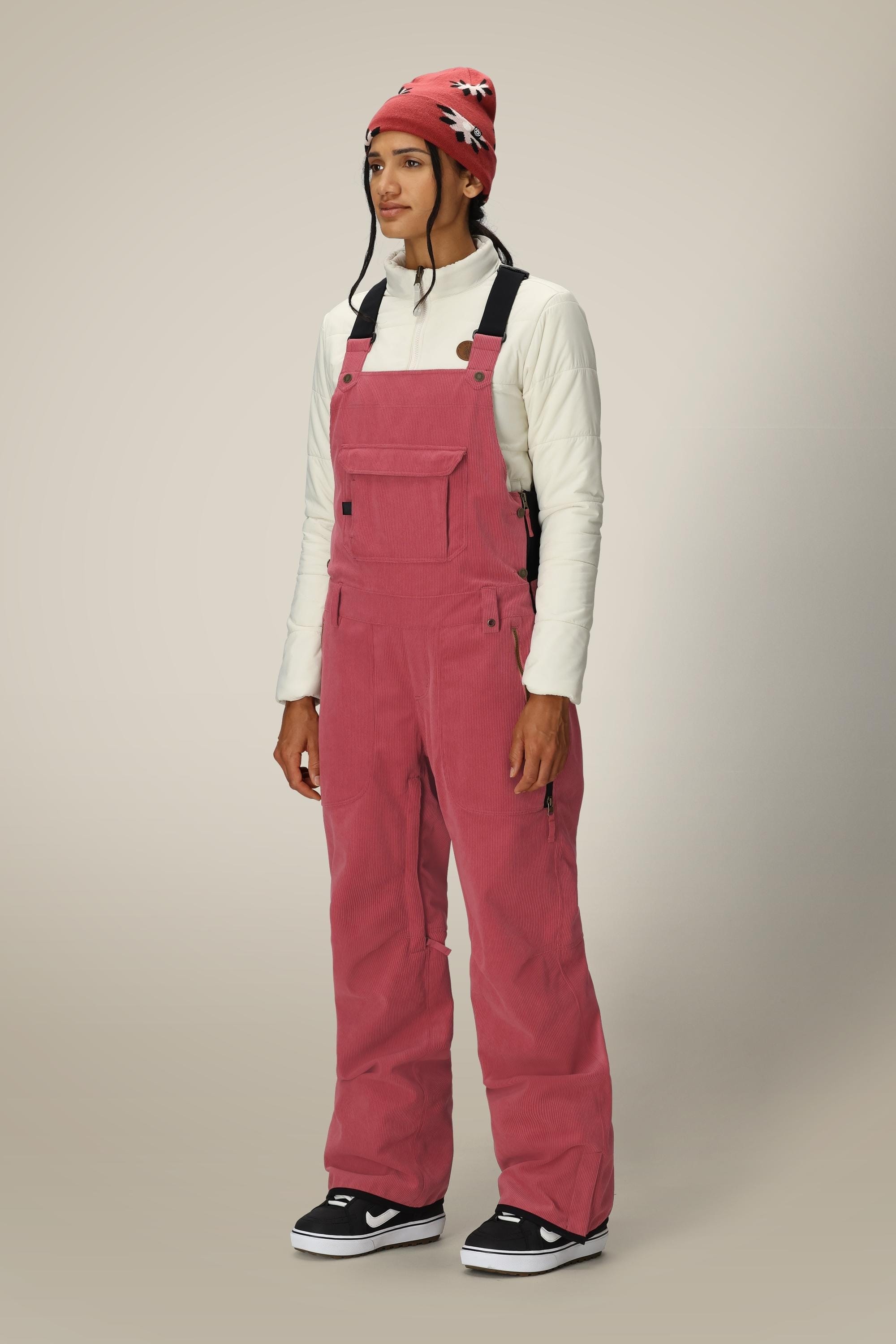 A person stands wearing a white long-sleeve top, the 686 Women's Black Magic™ Insulated Bib by 686, black-and-white sneakers, and a red beanie with a white and black pattern, against a plain light background.