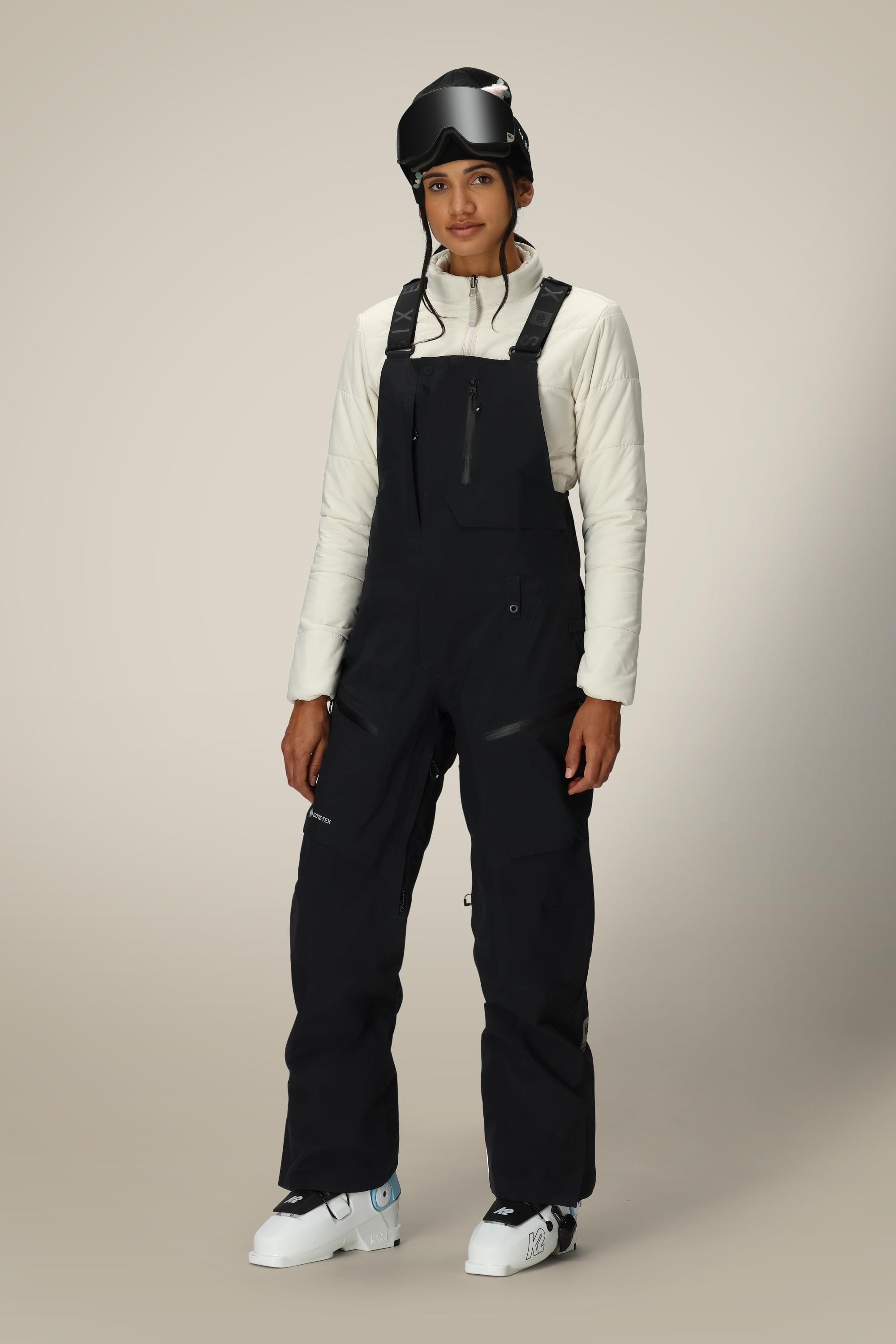 A person in a white long-sleeve top, 686 Women's GORE-TEX Dispatch™ Shell Bib ski pants, ski boots, and a black helmet with goggles stands against a plain light background.
