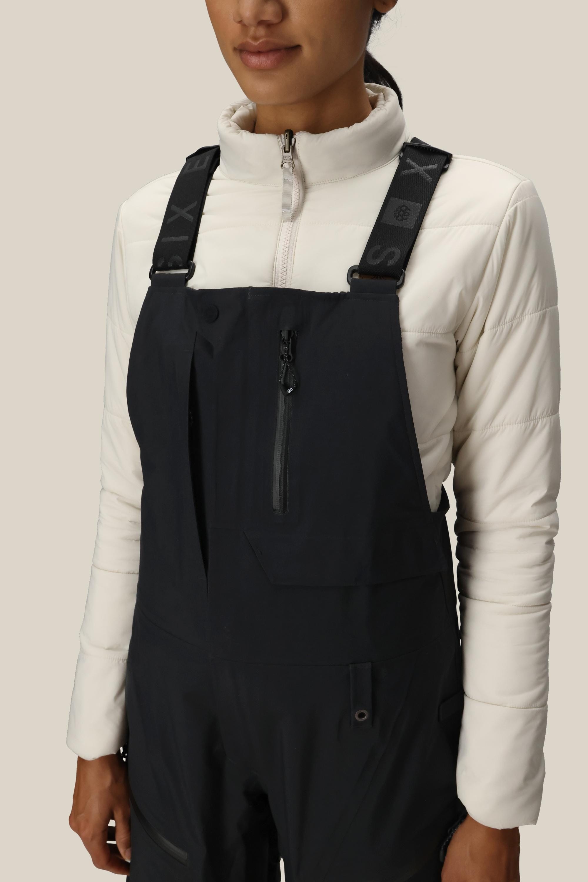A woman wearing the 686 Women's GORE-TEX Dispatch™ Shell Bib with adjustable straps and a white quilted long-sleeve top stands against a plain beige background.