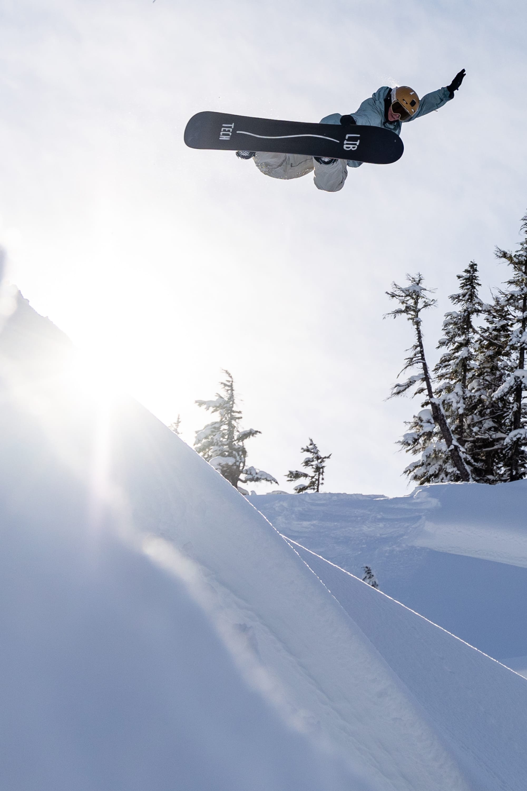 A snowboarder wearing the 686 Women's GORE-TEX Dispatch™ Shell Bib and a helmet catches air above a snowy slope, with sunlight beaming and evergreens in the background.