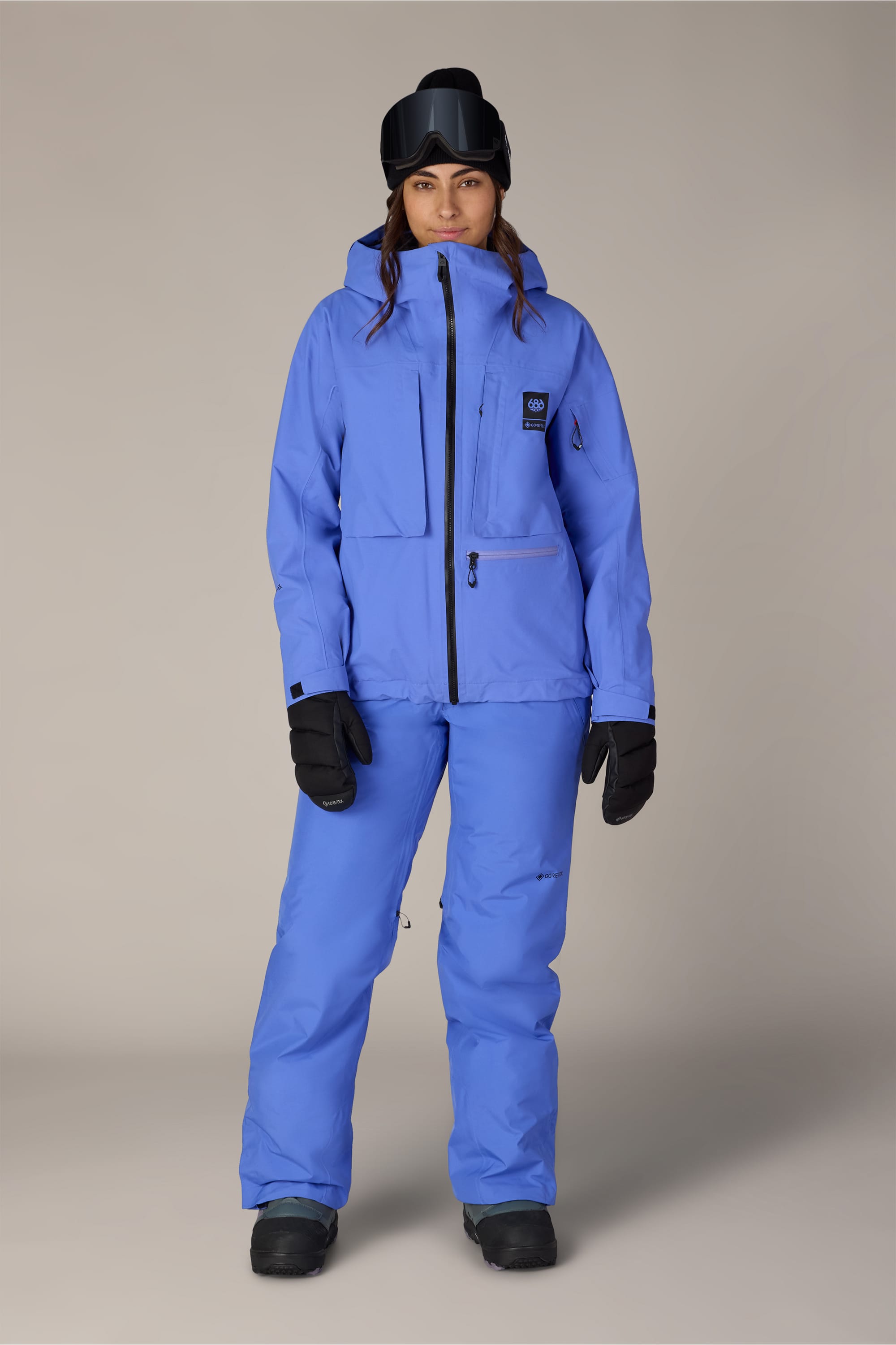 A person stands facing forward against a plain light gray background, wearing the 686 Women’s GORE-TEX® Willow™ Insulated Pant, paired with a matching light blue ski jacket, black gloves, black boots, and a black ski mask with goggles.