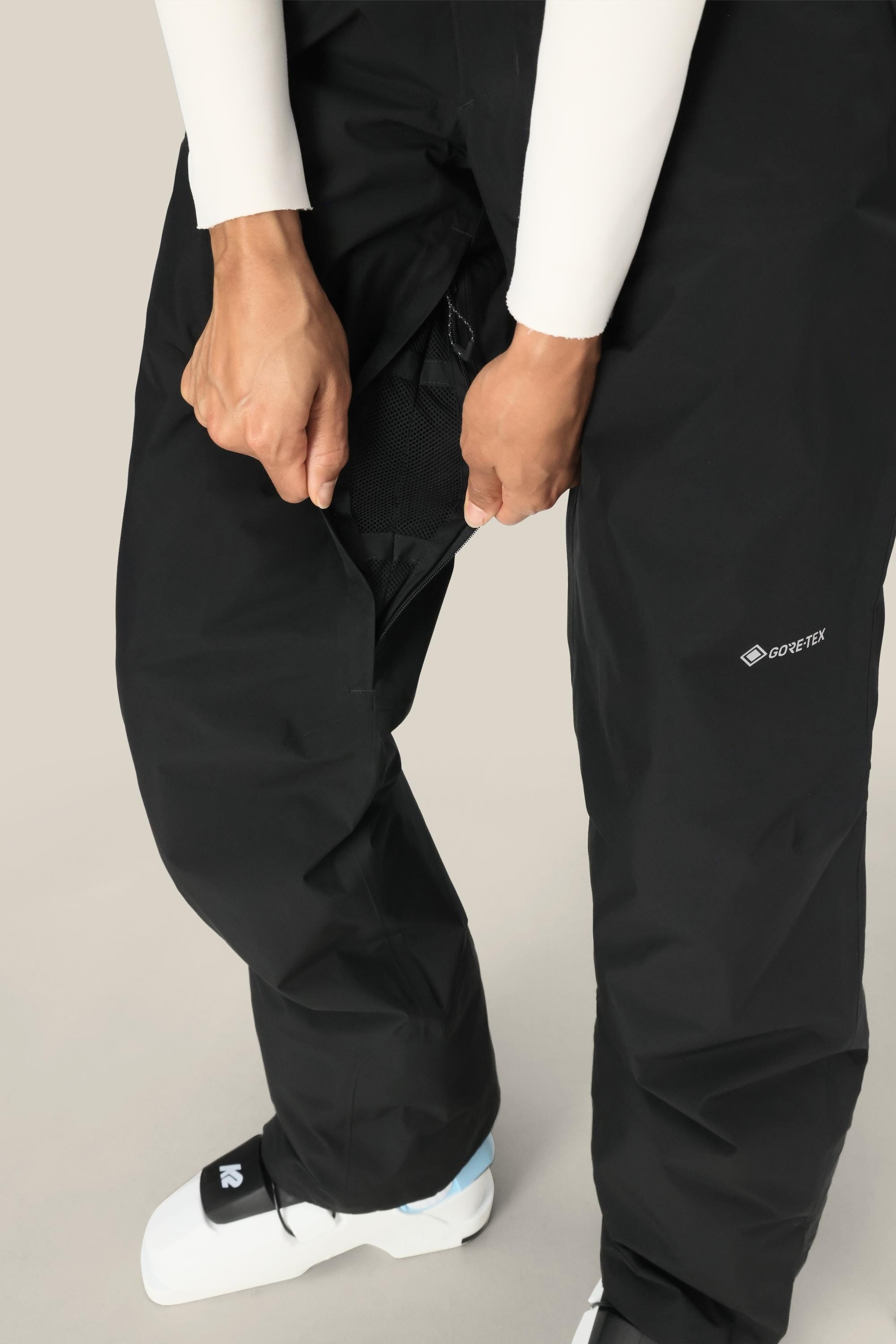 A person in 686 Women's GORE-TEX Willow™ Insulated Pants unzips the side to show the mesh lining, paired with white ski boots. The background is plain and light-colored.