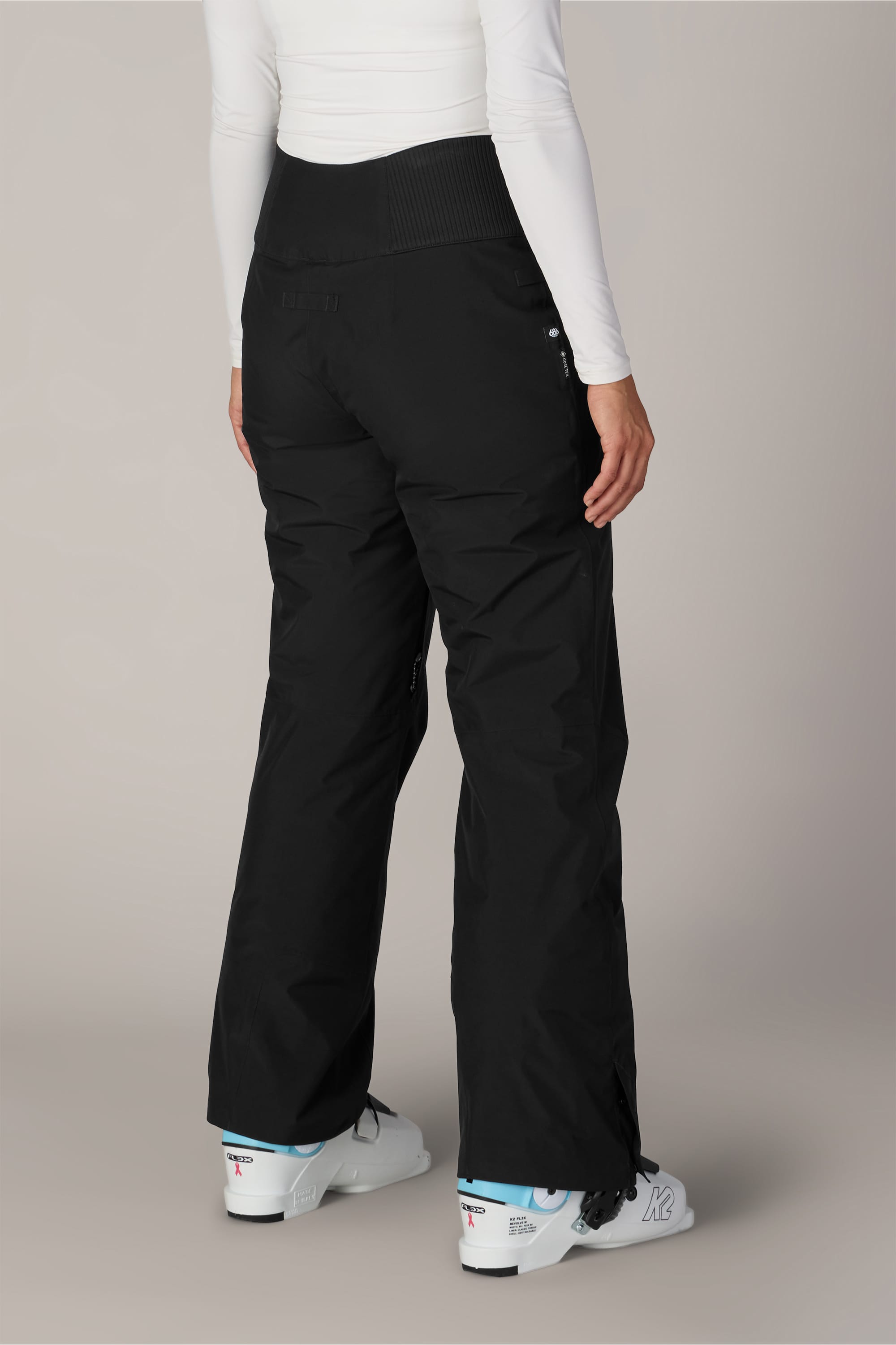 A person is shown from the lower back down wearing the 686 Women’s GORE-TEX® Willow™ Insulated Pant in black, paired with a white long-sleeve top and white ski boots, standing against a plain background.