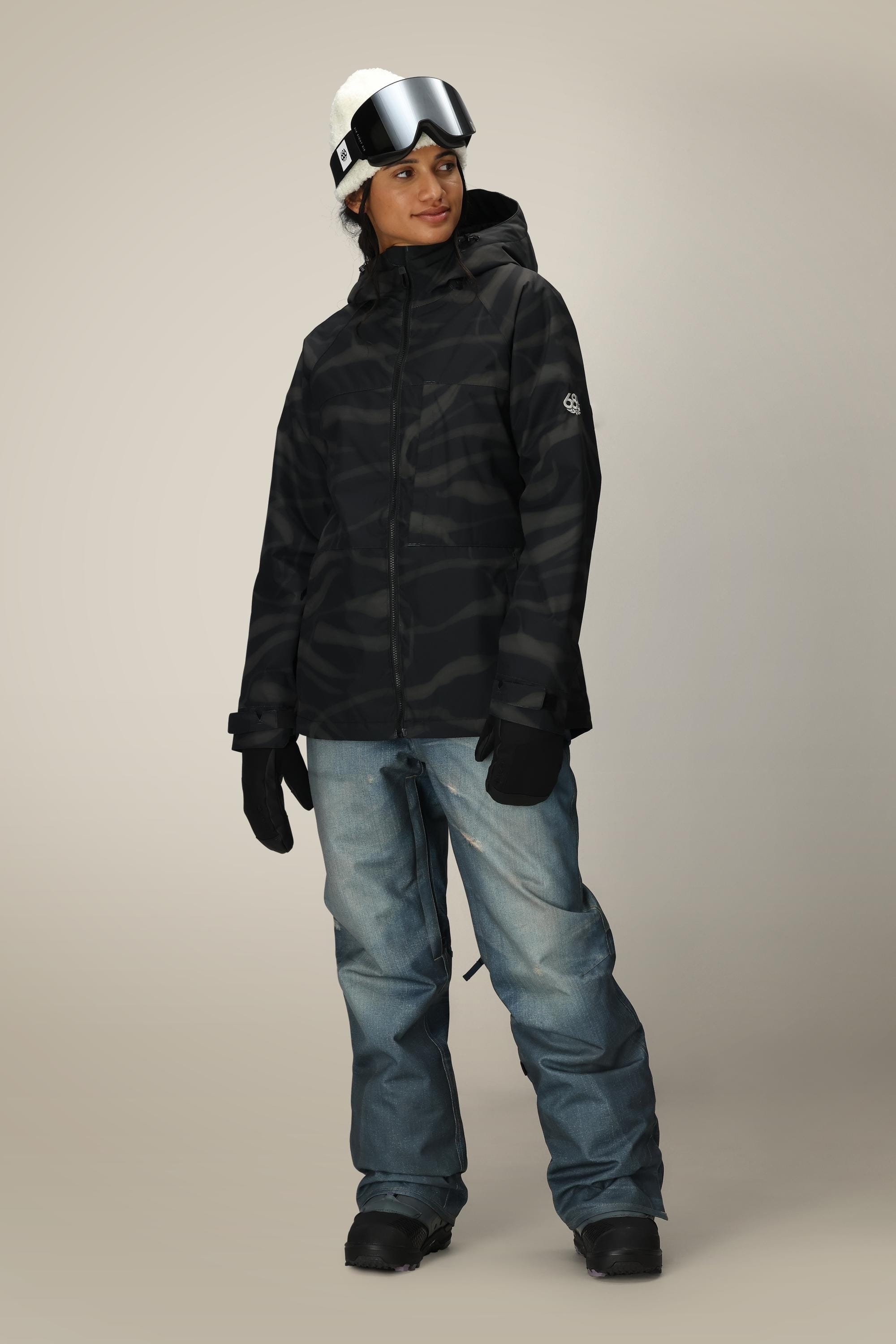 A person in a black camouflage winter jacket, 686 Women's Deconstructed™ Denim Insulated Pant by 686, black gloves and boots, white beanie, and ski goggles stands against a plain light background.