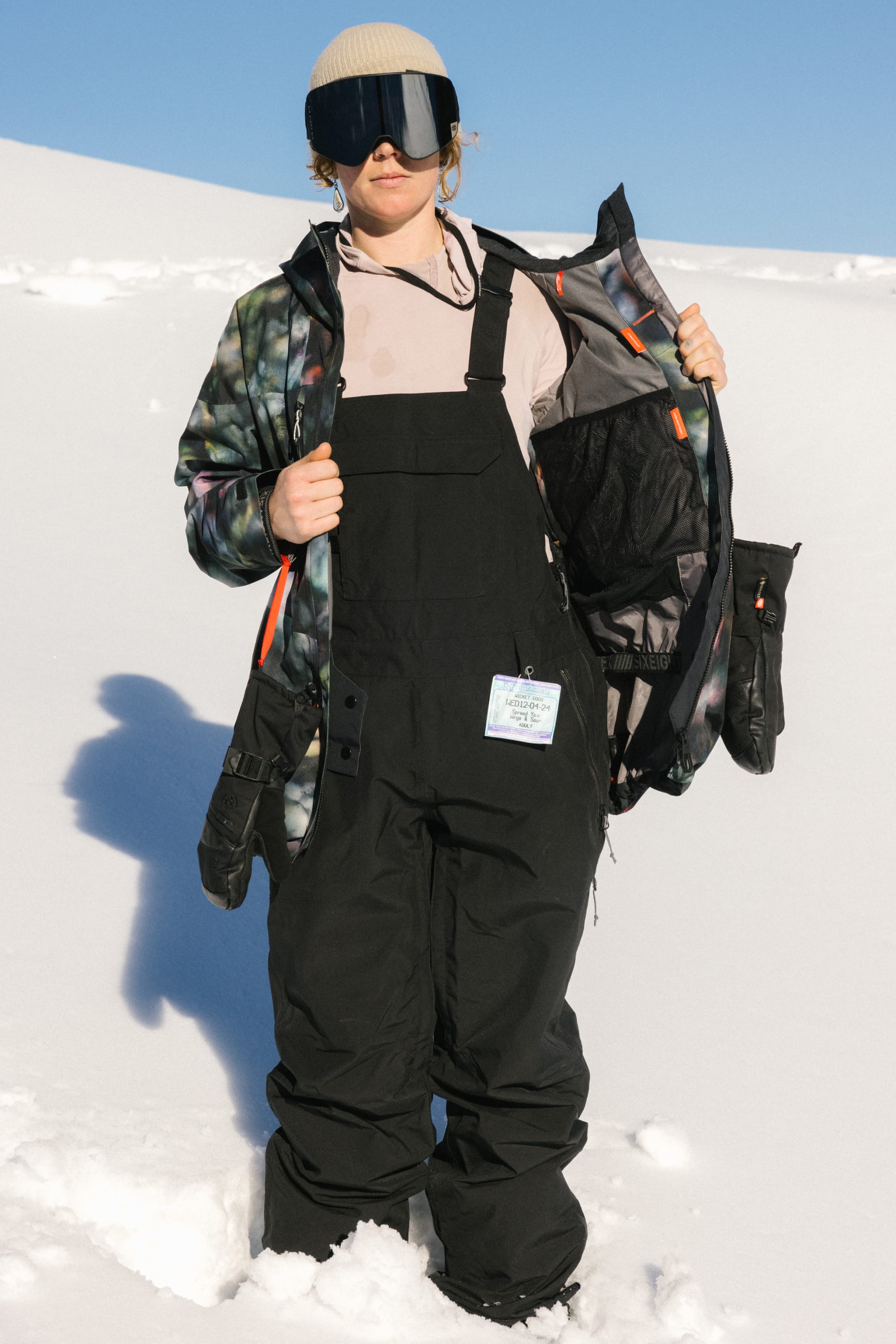 A woman wearing the 686 Women's Black Magic™ 20K Shell Bib stands on snow in a patterned jacket, goggles, and beige beanie, holding her jacket open to reveal the lining beneath a clear blue sky.