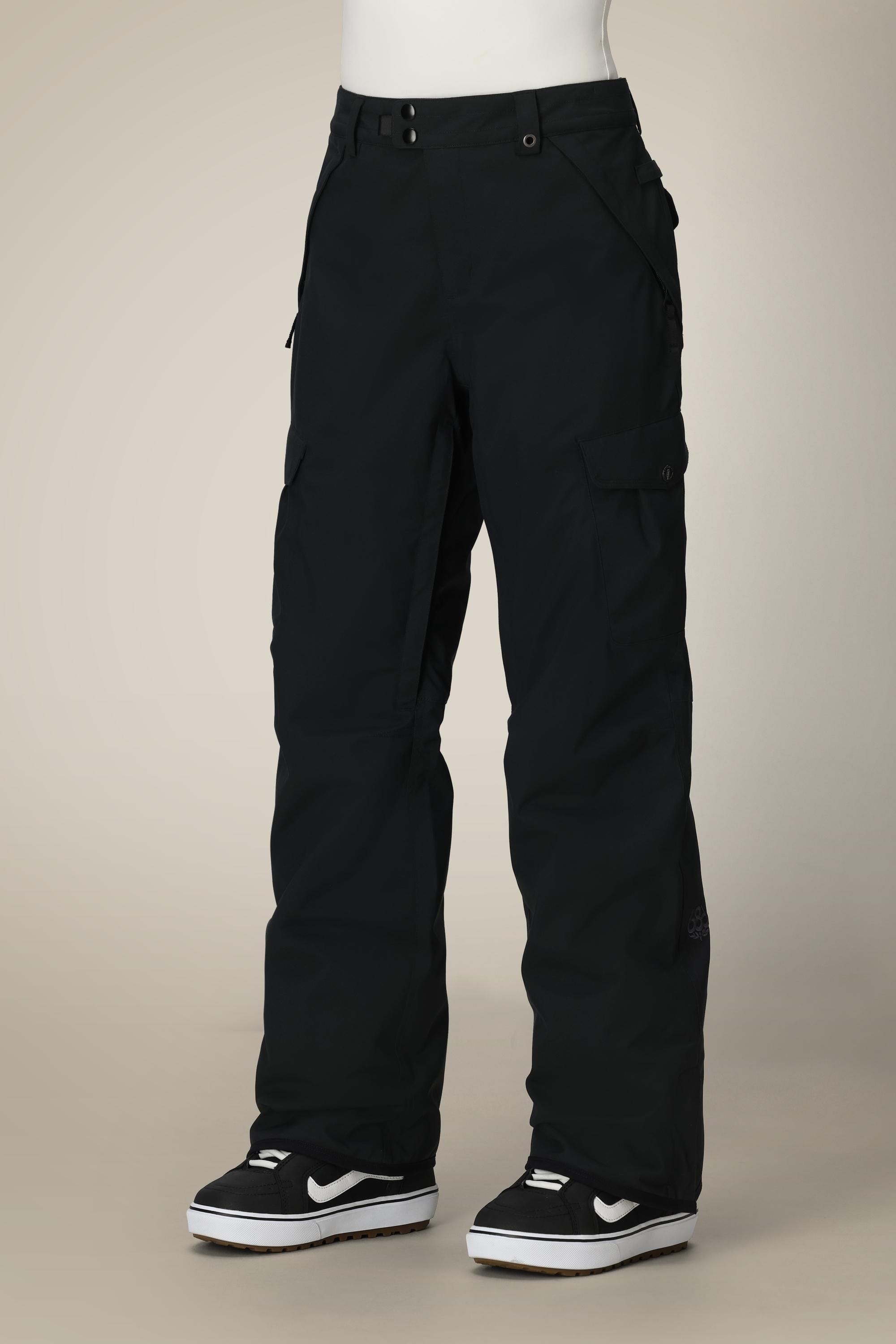 A person wears 686 Women's Aura™ Insulated Cargo Pant by 686—black, waterproof, relaxed-fit with multiple pockets—paired with black-and-white sneakers, standing against a beige background.