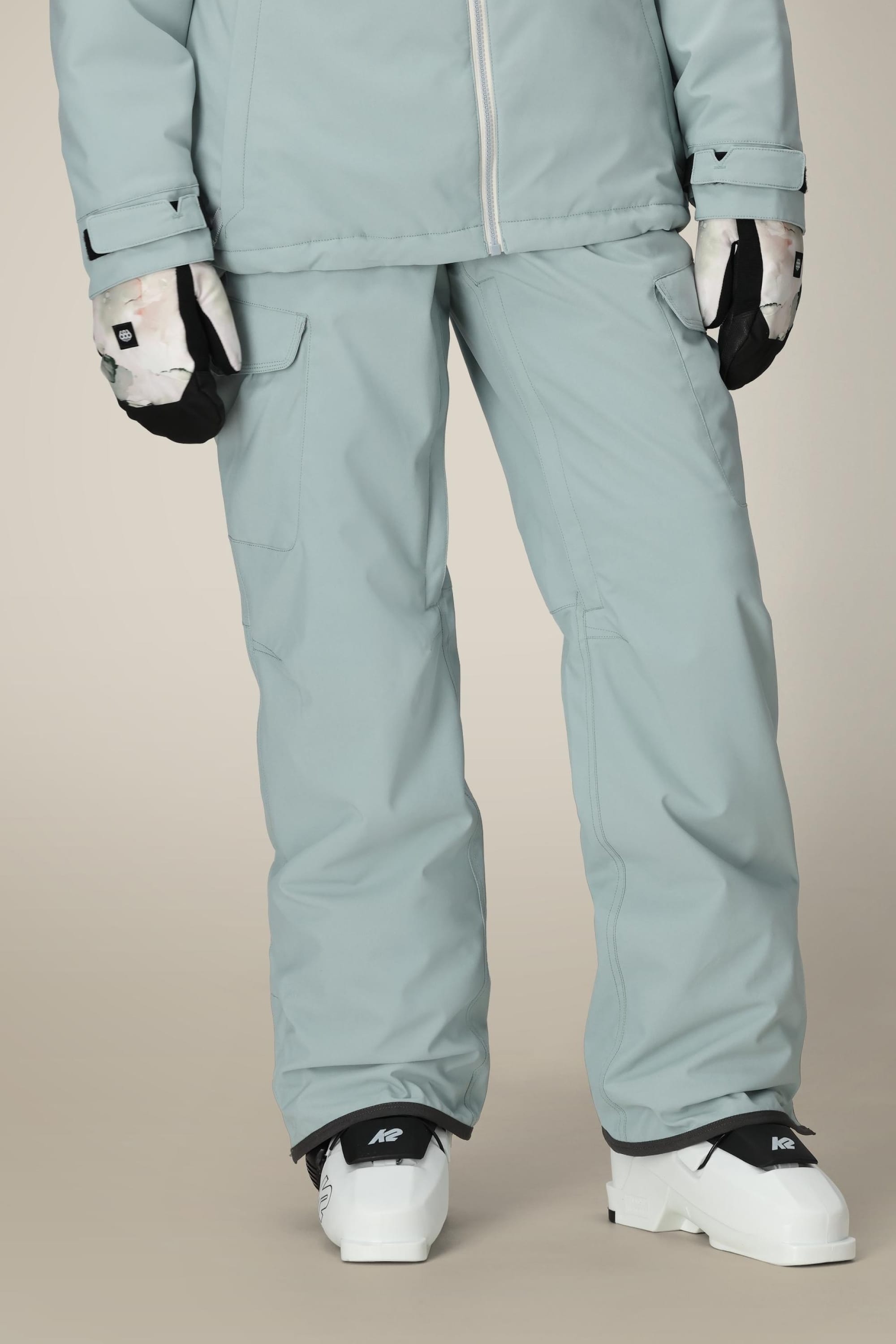 A person wears 686 Women's Aura™ Insulated Cargo Pant in light gray, a matching jacket, white and black gloves, and white ski boots while standing against a neutral background.