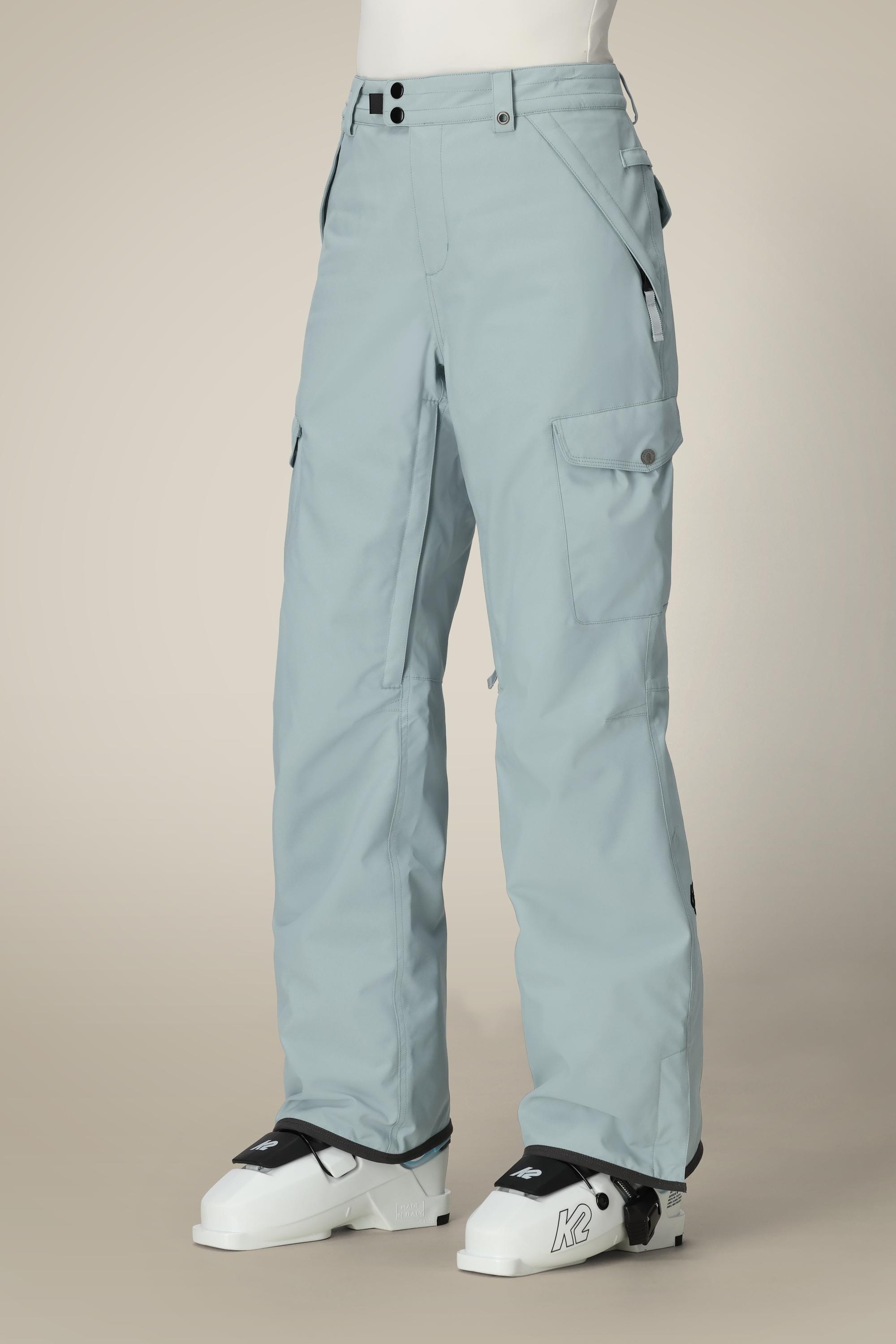 Person wearing 686 Women's Aura™ Insulated Cargo Pant in light blue, featuring waterproof fabric, multiple pockets, and a relaxed fit, paired with white ski boots with K2 branding against a neutral background.