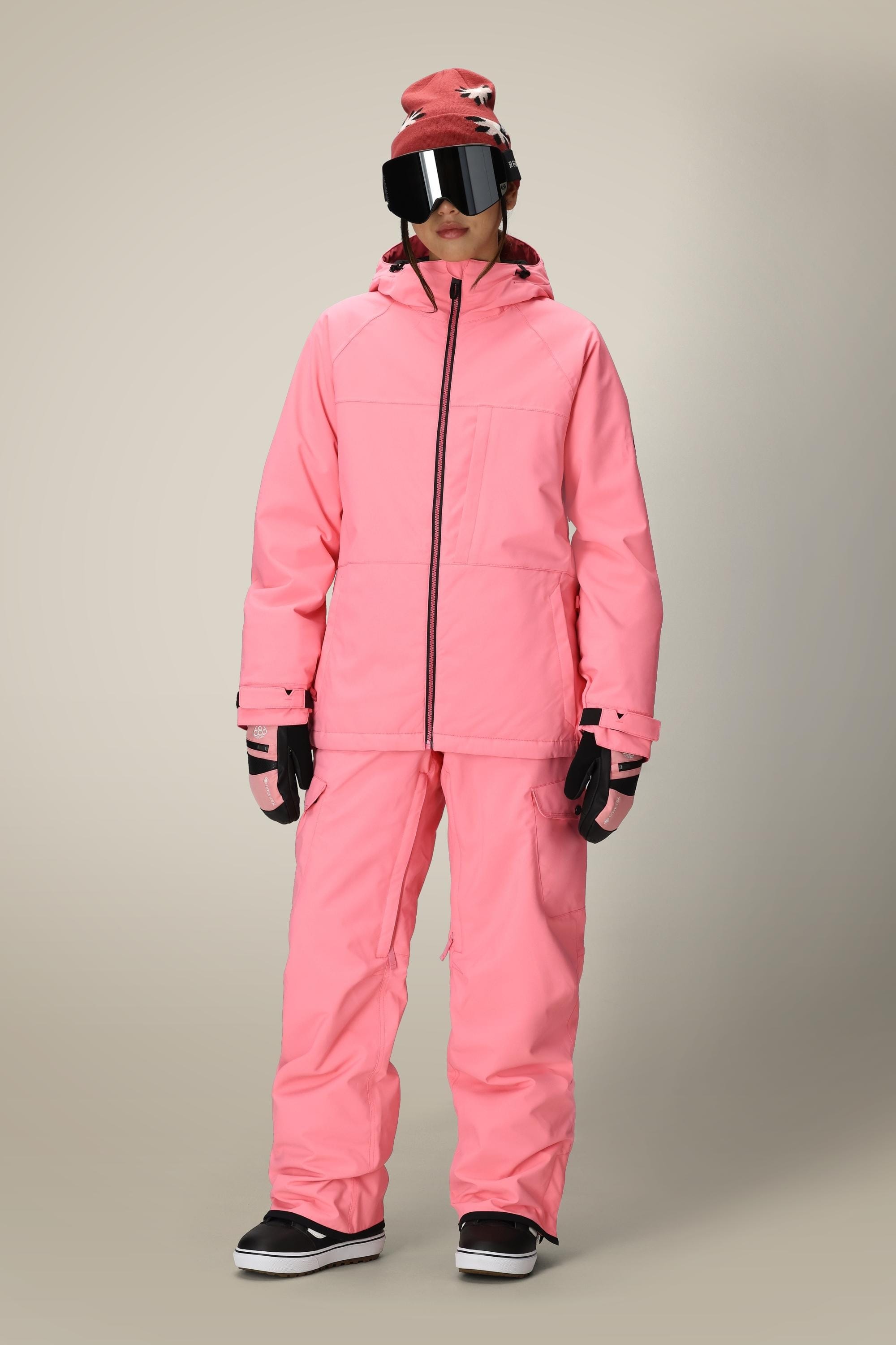 A person wearing a bright pink winter jacket, 686 Women's Aura™ Insulated Cargo Pant by 686, black gloves and shoes, a pink beanie with a white pattern, and large reflective ski goggles stands against a plain background.