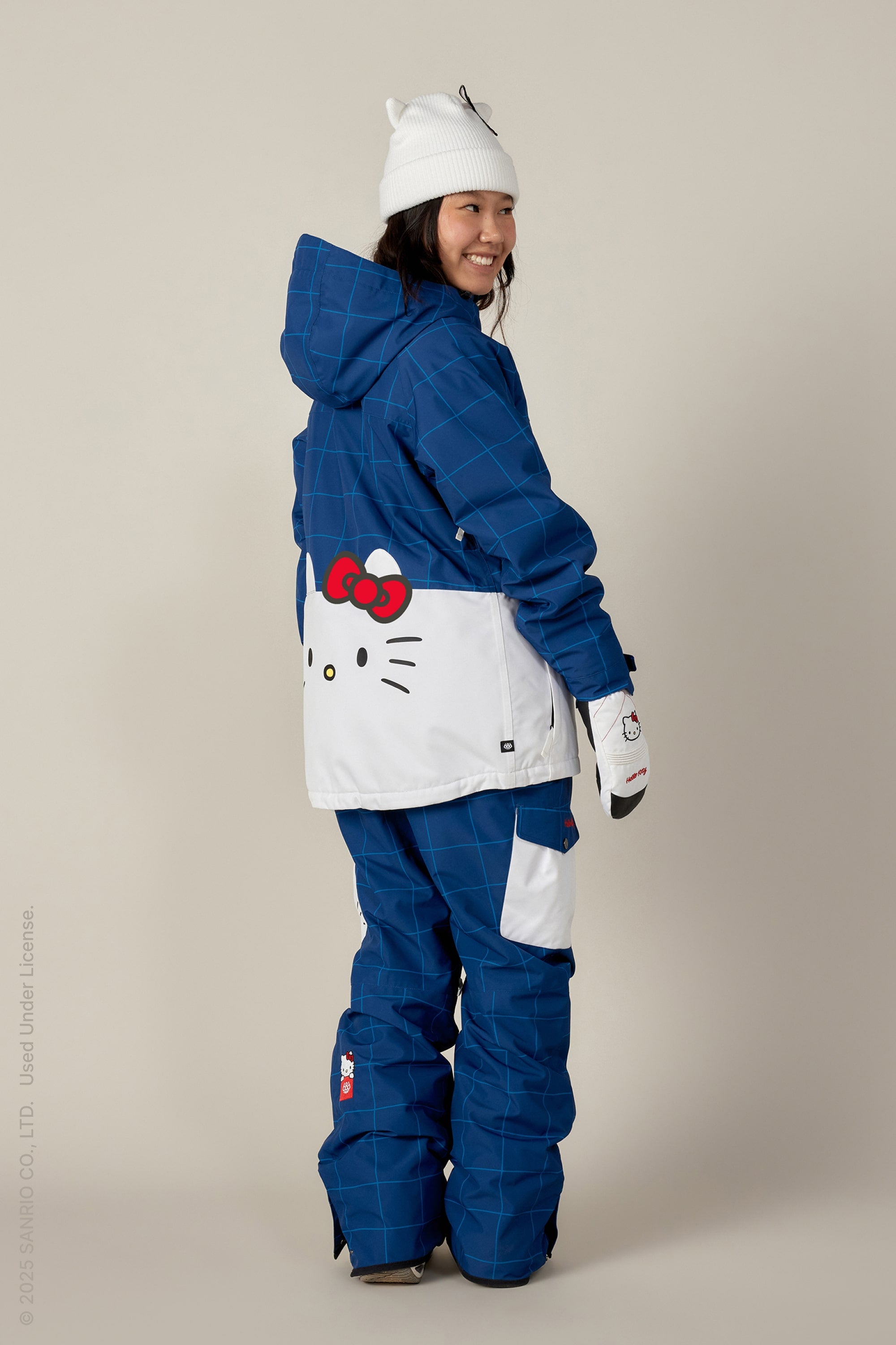 Wearing 686 Women's Aura™ Insulated Cargo Pants, a person dressed in a blue and white snow outfit with Hello Kitty details, white gloves, and a beanie stands smiling and looking over their shoulder.